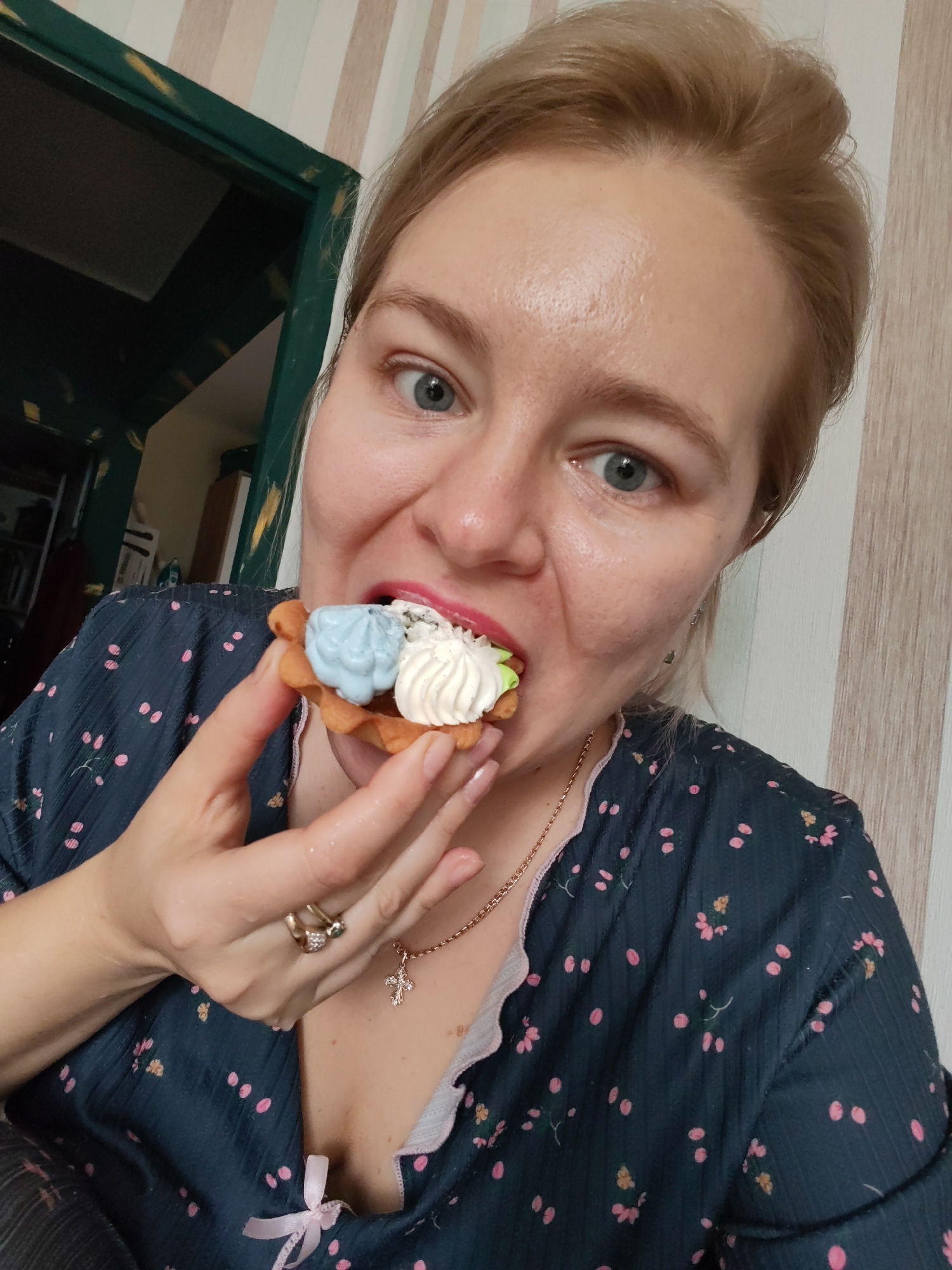 In a medium shot, a woman with fair skin and blonde hair pulled back is seen from the chest up, eating a small pastry topped with white and blue cream. She is wearing a dark blue patterned blouse with pink and yellow floral motifs and a scalloped trim. A delicate gold necklace with a cross pendant is visible around her neck, and she wears two rings on her left hand. The background consists of a wall with vertical stripes in cream and light brown, and a glimpse of a dark green doorway is visible to the left, suggesting an indoor domestic setting. The lighting is even, and the image is in focus, capturing a casual moment.