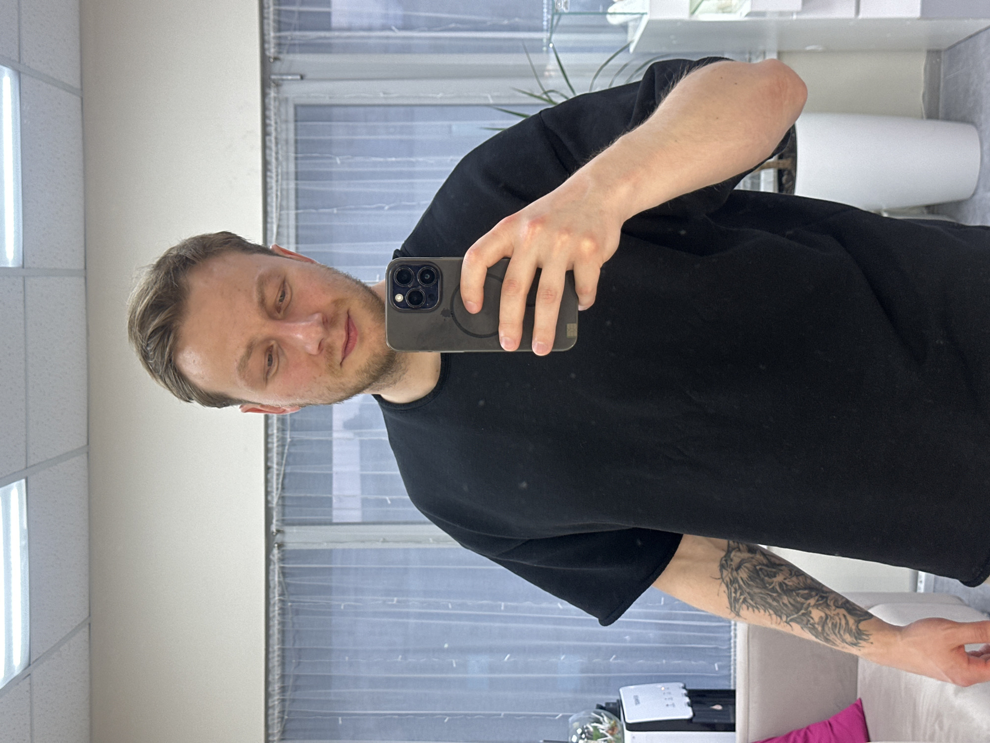 An upright mirror selfie shows a man of average build, with short light brown hair and light stubble, looking towards his smartphone screen with a neutral expression, hinting at a faint smile. He wears a plain black short-sleeved t-shirt. A prominent, detailed tattoo depicting what appears to be a wolf or similar animal head with intricate fur patterns covers a significant portion of his left forearm.
He holds a dark-colored iPhone, likely a Pro model with a triple camera array, encased in a clear cover that reveals a circular MagSafe charging ring on its back. The image is taken in a full-length mirror, reflecting the indoor setting.
The background features a large window with horizontal blinds or textured glass, and overhead, rectangular recessed LED light panels are visible on the ceiling. Reflected elements include a plant in a white cylindrical pot, some glass shelving, and parts of a light-colored sofa or armchair in the lower right, with a bright pink object or fabric visible behind it. The environment appears clean, modern, and well-lit, consistent with an office, salon, or contemporary apartment in Sochi, Russia. No visible text is discernible in the image.