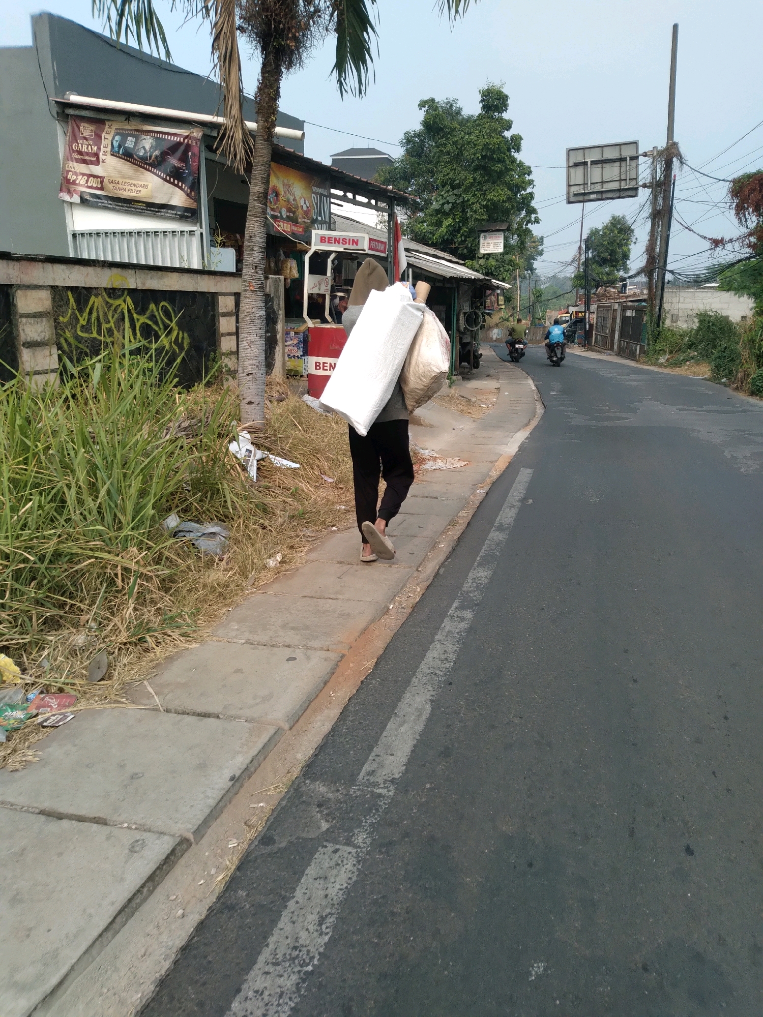 In South Tangerang, Indonesia, a person walks along a concrete sidewalk next to a paved road. The person, viewed from behind, carries a large, white, bagged object over their shoulder. They wear dark pants and light-colored sandals. To the left of the sidewalk is overgrown dry grass with scattered debris. Adjacent to the grass is a low concrete wall, some parts of which are covered in graffiti. Along the road, a few motorcycles are visible; one is ridden by a person wearing a blue shirt. Buildings line the street, including one with a banner displaying "GUDANG GARAM INTERNATIONAL" and another with "BENSI N". Overhead, utility poles with numerous wires stretch across the scene, and a large, empty billboard is mounted on a pole. The sky is overcast.