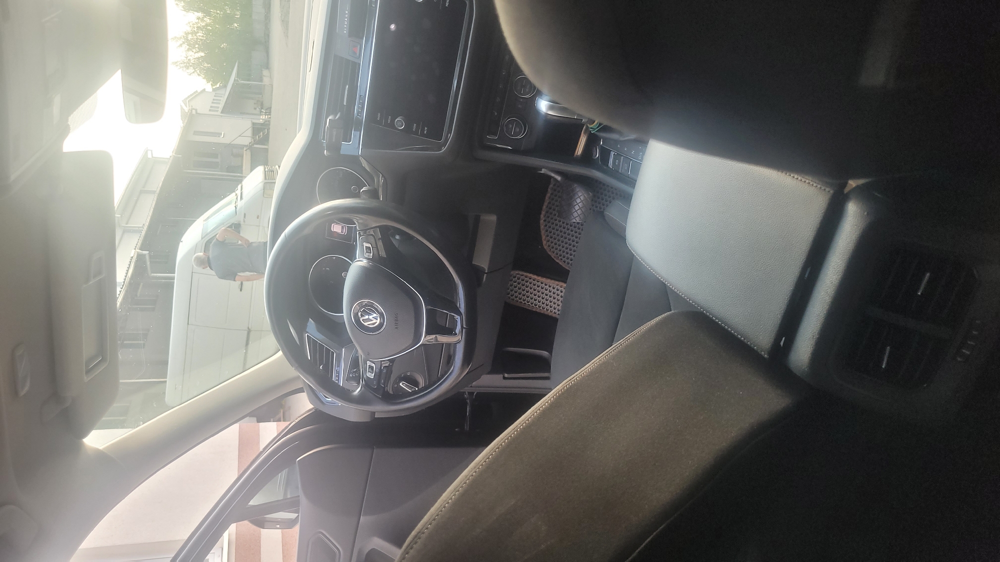 The image shows the interior of a Volkswagen car, focusing on the steering wheel, dashboard, and center console. A person is visible through the windshield, standing next to a white van outside the car. The scene appears to be an outdoor environment, possibly a parking lot, with buildings and trees in the background. The lighting suggests it is daytime, with bright sunlight.