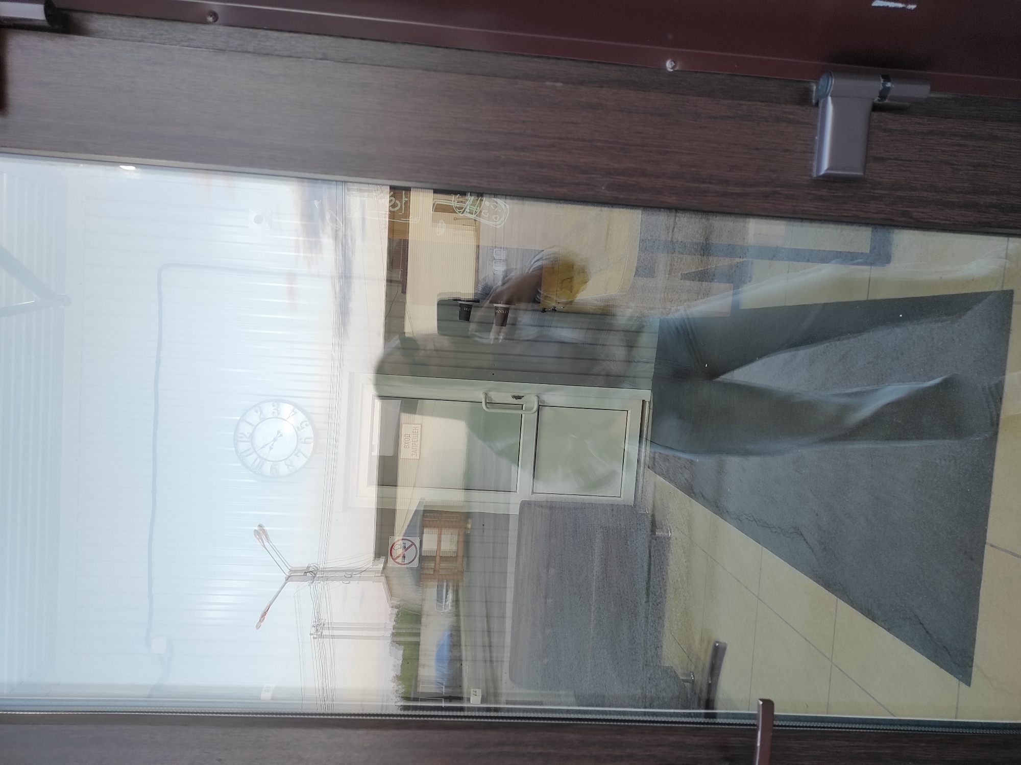 A photograph captures a complex reflection in a large glass pane, framed by a dark brown, wood-patterned border. The image is oriented vertically, showing the glass pane extending from left to right.
Through the glass, an exterior scene is visible: a tall, multi-limbed utility pole or structure with numerous power lines stretching across a light, possibly overcast sky. Distant, low-rise buildings can be vaguely discerned beyond.
The glass heavily reflects an interior space. A person's silhouette, likely holding a smartphone, is prominent in the reflection, suggesting they are taking the photo. Reflected inside is another white-framed glass door with a handle, beyond which a tiled floor is visible. A dark gray floor mat is positioned before this inner door. Above the inner door, a round wall clock with Roman numerals is reflected, showing hands pointing towards approximately 9:50 or 8:50.
Notable details in the reflection include a white sign on the inner door that clearly states "BXOД ЗАПРЕЩЕН!" (Entry Forbidden!). Below this, a red circular "No Smoking" symbol is also reflected. Some indistinct objects appear on what might be a counter or shelf within the reflected interior. A brown-colored latch or door stopper mechanism is attached to the top-right part of the outer door frame.
The overall scene is a daytime shot, likely taken indoors looking out through a glass door or window in a commercial or public building in Chaikovsky, Russia. The light is diffused, suggesting an overcast day or indirect light.