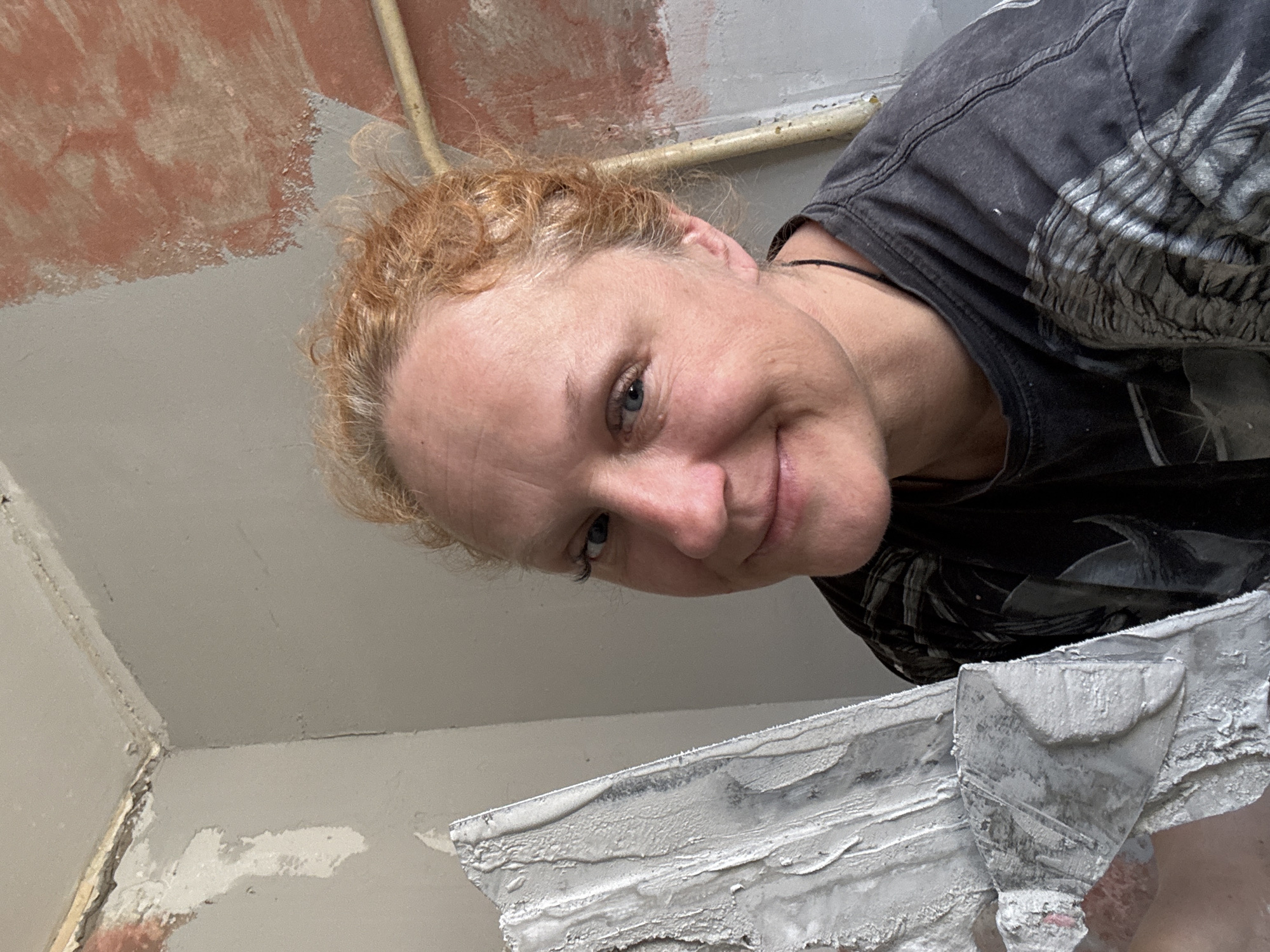 A woman with red-blonde, curly hair is positioned in the foreground of the image, looking down and smiling slightly. She is wearing a dark-colored t-shirt with a faded design. In front of her, a metal trowel coated in white plaster or spackle is visible. The background shows an unfinished or partially renovated interior, featuring walls and a ceiling. The walls appear to be painted with a light gray, while sections of the ceiling show a reddish-brown color, suggesting an older layer of paint. A light-colored pipe runs along the ceiling. The exact location in the city of Chaikovsky, Russia, cannot be determined from the visual information provided.