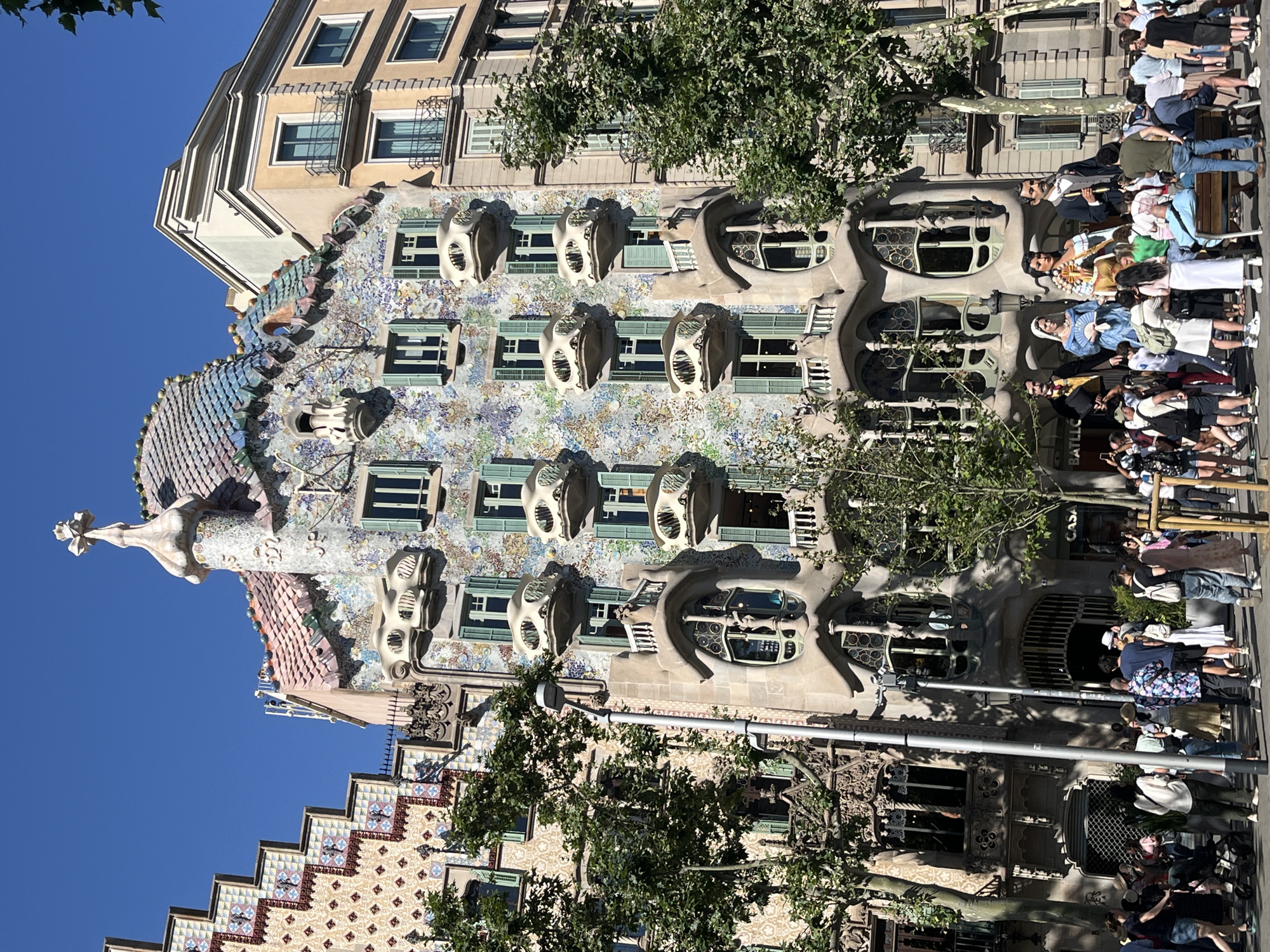 The image shows a street view of Casa Batlló in Barcelona, Spain, a distinctive building designed by Antoni Gaudí. The facade is characterized by its colorful, mosaic-like tiling, organic shapes, and unique balconies that resemble skulls. Several large, festive figures, known as "gigantes" or giants, are positioned in front of the building. A crowd of tourists and locals are gathered on the sidewalk, some taking pictures of the building and the figures. Trees line the street, and the sky is clear and blue. The architectural style is a clear example of Catalan Modernism.