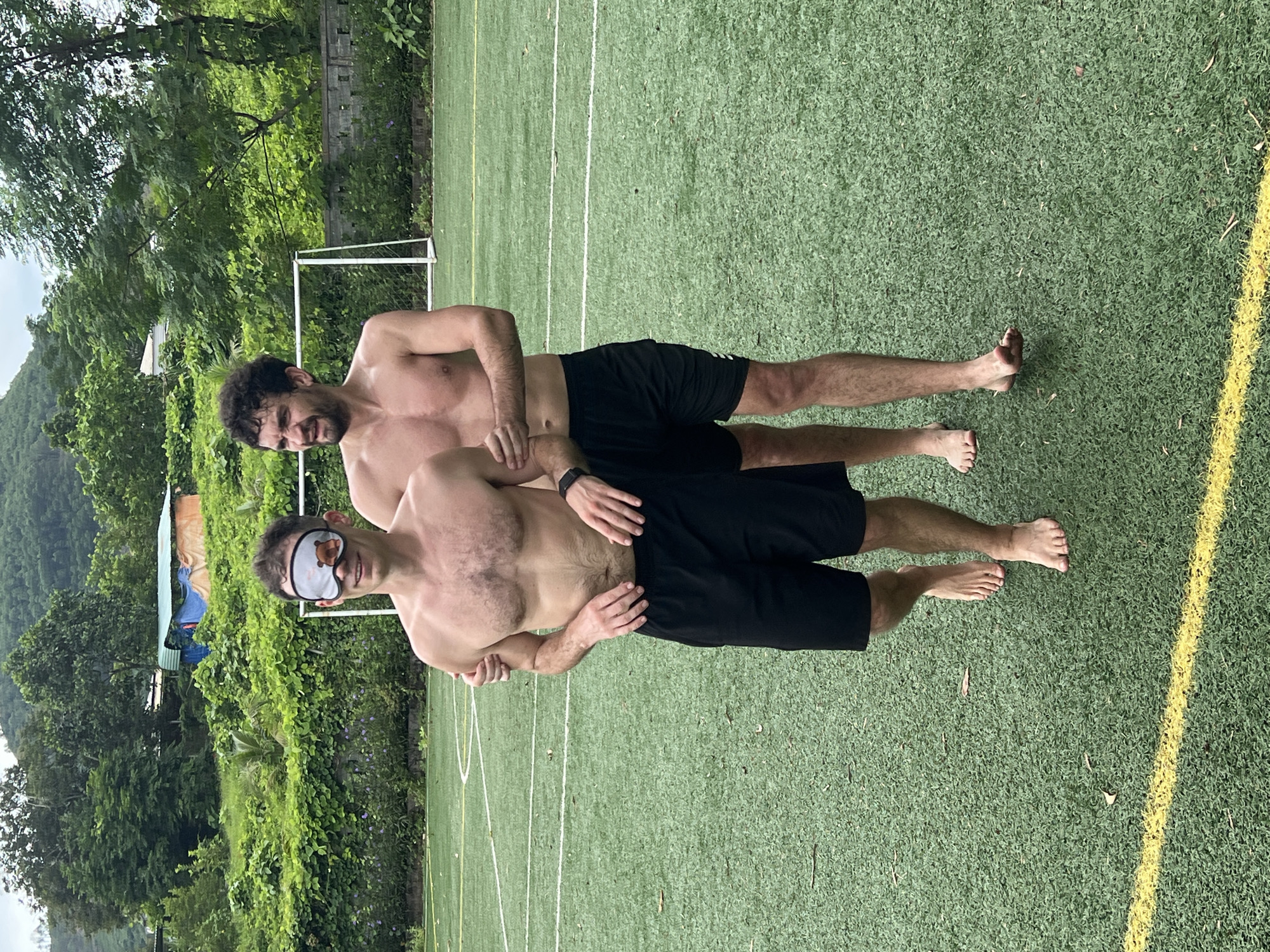 Two shirtless men stand on a green turf field, possibly a soccer field, in Đà Nẵng, Vietnam. The man in the foreground wears a blindfold with a teddy bear design over his eyes and black shorts. He is barefoot and appears to be walking. The man behind him, also wearing black shorts, has his arms around the first man's shoulders. He is looking towards the left with a slight grimace. The background shows lush green vegetation and trees, suggesting a natural or park-like setting within the city. The sky is overcast.
