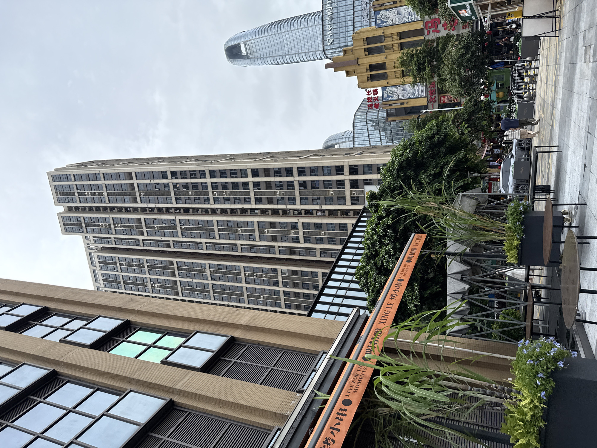 The image depicts a street scene in Haizhu District, China, characterized by a collection of modern high-rise buildings. In the foreground, a light brown building with horizontal louvered windows and rows of square windows is on the left. An orange awning with Chinese characters and English text, "LIVE FOR THE MOMENTS XING LE," is visible extending from this building, partially obscuring a metal support structure. Several tall, reedy plants and potted greenery with small blue flowers are placed in front of the awning, creating a cafe-like setting with tables and chairs.
Dominating the center of the image is a very tall, slender skyscraper with a grid of numerous windows. To the right of this, another tall, curved skyscraper with a distinctive cutout at the top is visible, along with other modern glass-fronted buildings and a yellow building with architectural detailing. The ground is paved with light-colored square tiles, and a sidewalk runs along the right side of the street.
In the middle ground, a gray sedan is parked on the street. Two individuals, a person in a light-colored striped outfit and another in a dark blue shirt and pants, are walking away from the camera on the sidewalk. Further down the street, more people are visible, along with storefronts and trees lining the walkway. The sky is overcast and gray. The scene suggests an urban commercial or residential area.