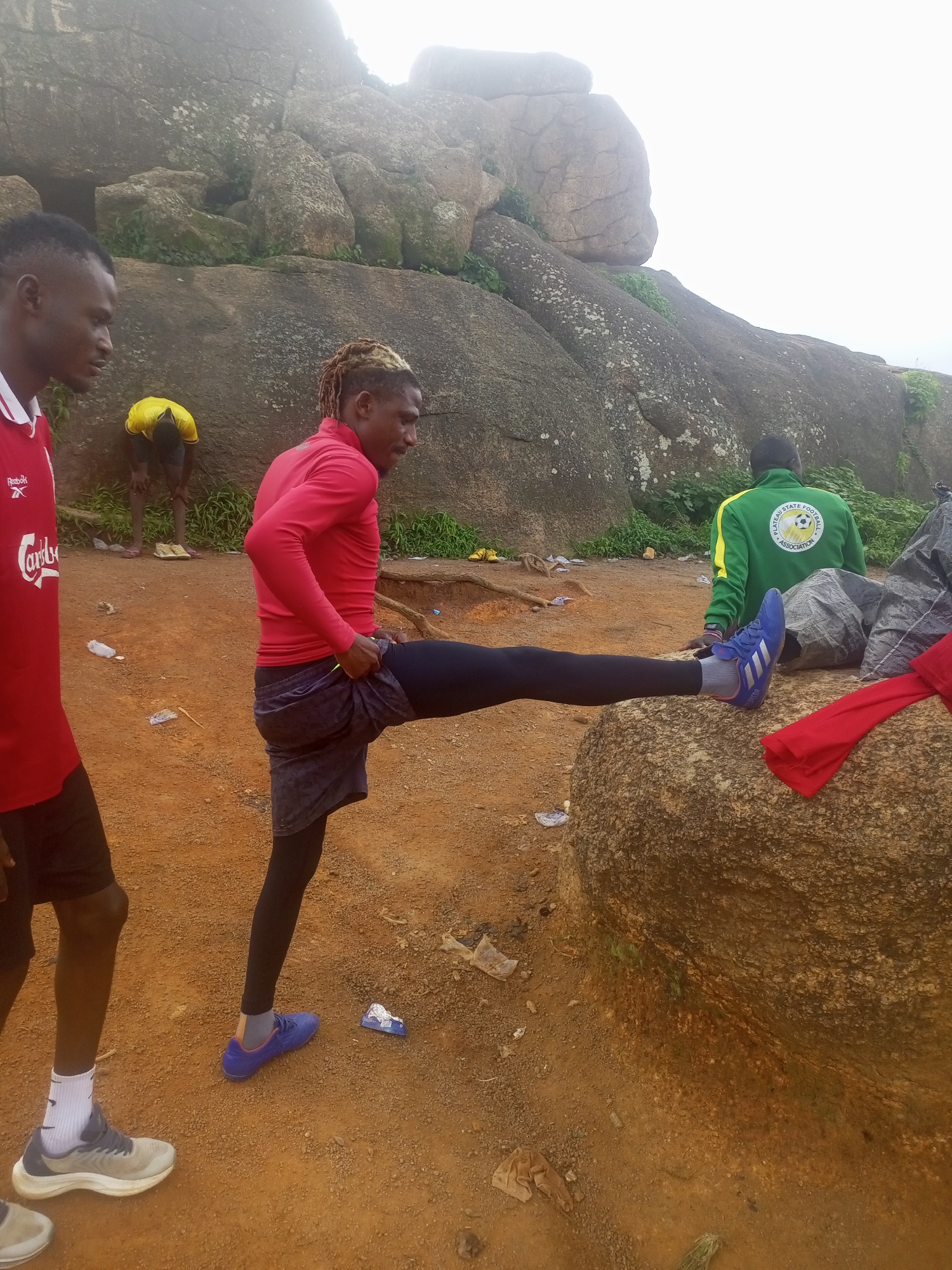 The image depicts a group of young men in an outdoor setting, likely in the city of Jos, Nigeria, known for its rocky terrain. The primary subject is a man in a red long-sleeved shirt and dark shorts over black leggings, stretching his right leg out onto a large rock. He is wearing blue athletic shoes. To his left, another man in a red shirt with white lettering and black shorts stands partially in the frame. In the background, a man in a yellow shirt is crouched down, and a man wearing a green and yellow jacket with "PLATEAU STATE FOOTBALL ASSOCIATION" printed on the back is seated on the ground. The environment consists of reddish-brown dirt ground, scattered rocks, and sparse vegetation, with large rock formations dominating the background. The sky is overcast, suggesting a cloudy or hazy day. There are also various pieces of debris on the ground, including plastic wrappers and what appears to be fabric. The overall scene suggests an informal athletic or training activity taking place in a natural, rocky landscape typical of parts of Jos.