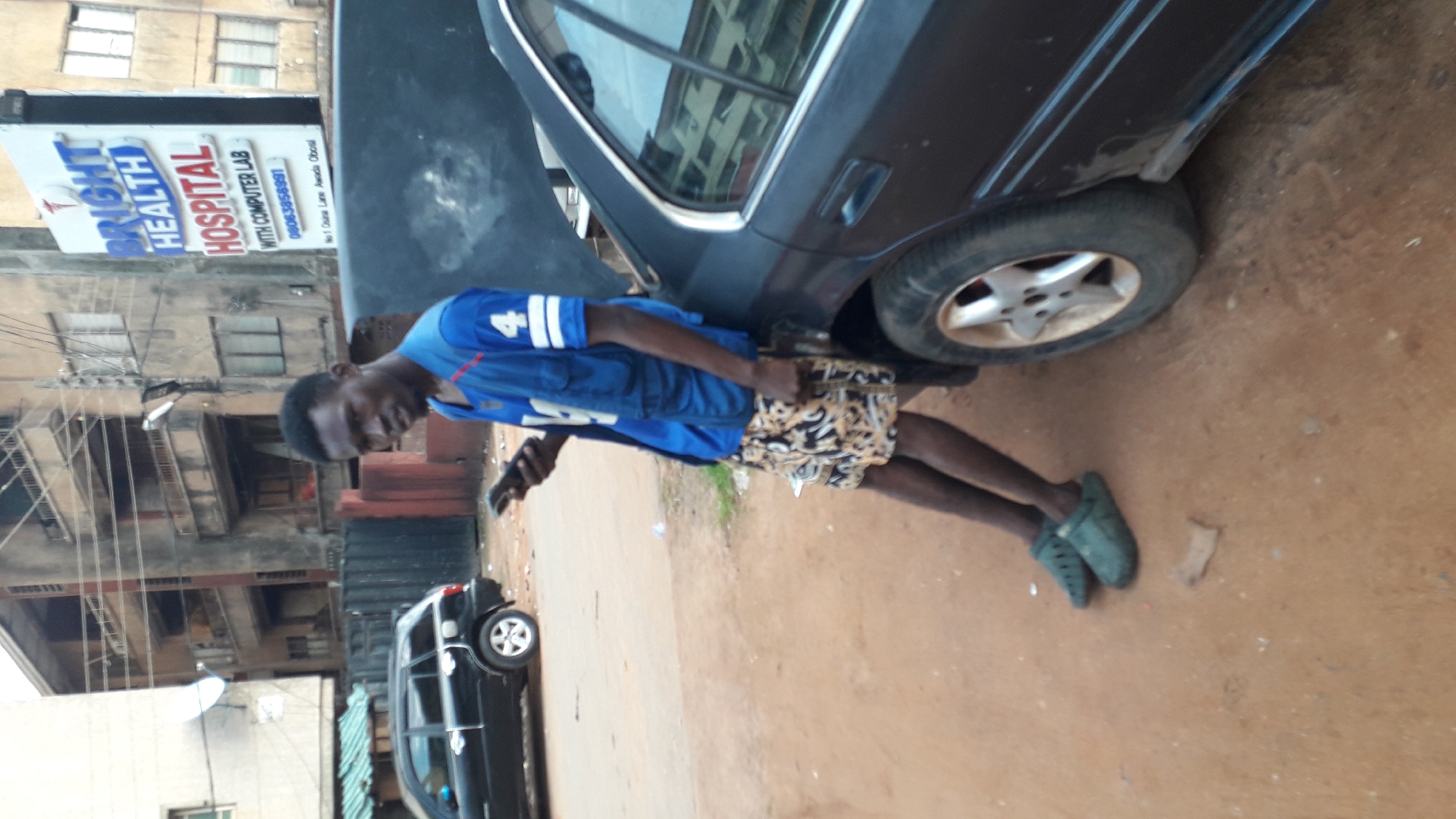 A young man in a blue and white jersey with the number "4", patterned shorts, and green shoes stands beside a dark-colored car. He is holding a mobile phone. Behind him, a sign reads "BRIGHT HEALTH HOSPITAL WITH COMPUTER LAB" and lists a phone number with the Nigerian country code. A black vehicle is visible further back in the scene. The environment appears to be an outdoor street setting, likely in the city of Onitsha, Nigeria.