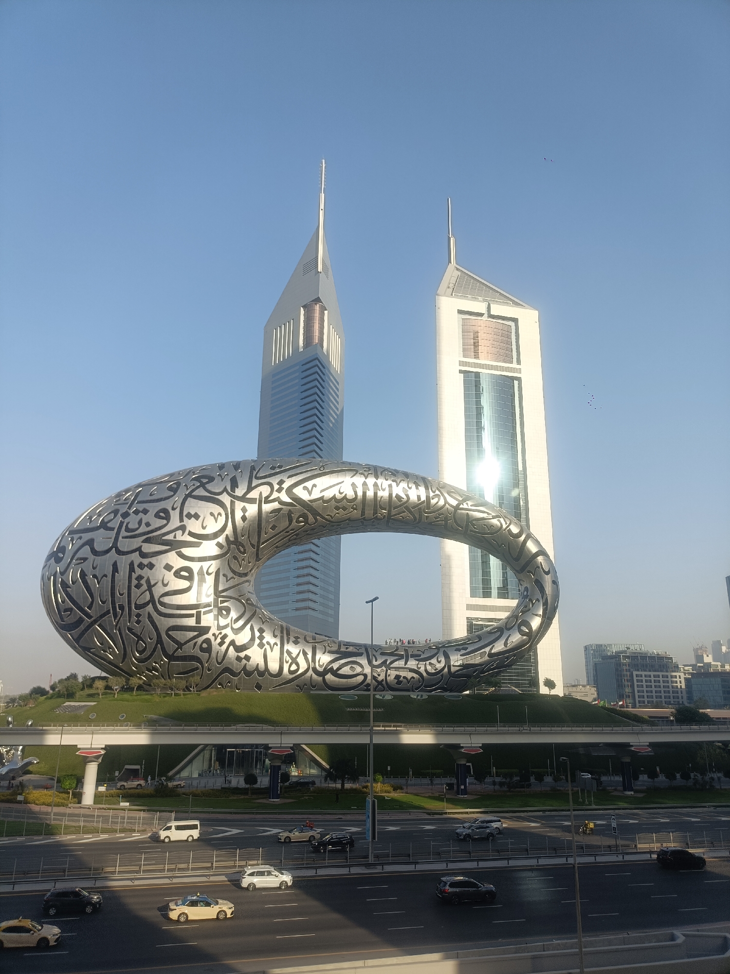 The image shows a street scene in Dubai, United Arab Emirates, with the Museum of the Future in the foreground and the Emirates Towers in the background. The Museum of the Future is a large, elliptical structure covered in Arabic calligraphy. The Emirates Towers are two tall, modern skyscrapers. Several cars and taxis are on the road in front of the museum. The sky is clear and blue. There are a few people visible standing on the Museum of the Future.