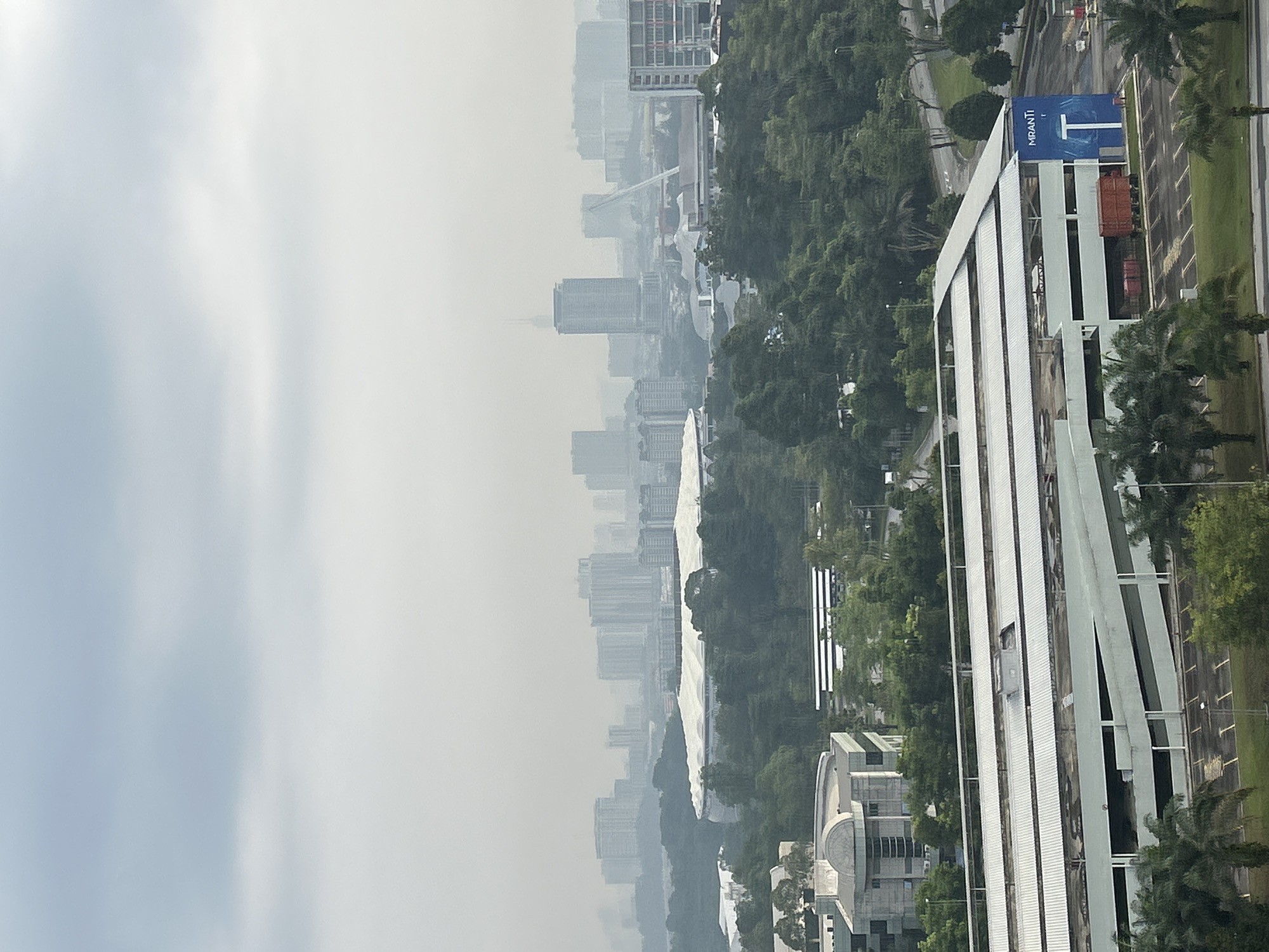 The image presents an elevated view of Kuala Lumpur, Malaysia, on what appears to be a hazy or smoggy day. The skyline is visible in the background, albeit obscured by the atmospheric conditions. Foreground elements include trees, a building with vertical white panels, and a parking area with a blue sign that reads "Mranti". Other buildings and roads can be seen in the middle ground. No people are visible in the frame.