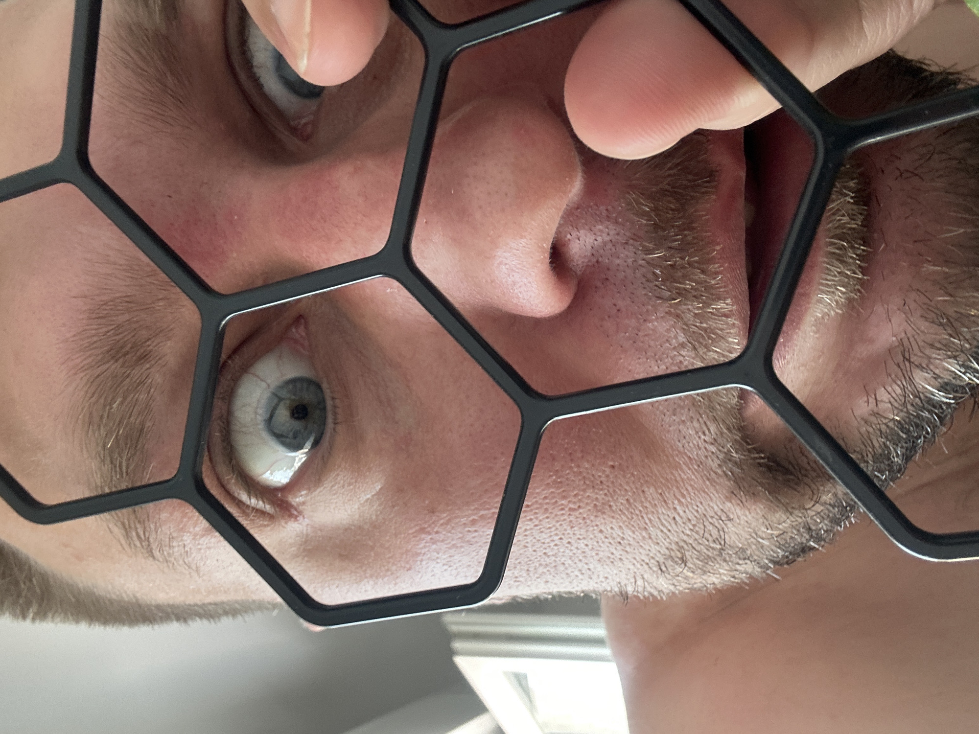 A close-up, low-angle shot captures a person's face as they peer through a black, hexagonal-shaped object. The subject, with light-colored eyes and short-cropped brown hair, exhibits a surprised or wide-eyed expression. Their left eye is visible, while the right eye is partially obscured by the object. A portion of the subject's nose and mouth are also visible, with a light beard stubble on their chin and upper lip. The hexagonal object appears to be a frame or grate, with a finger on the right side of the image appearing to hold it in place or adjust it. The background is out of focus, but suggests an indoor environment with a window and natural light on the left side. The location is not discernible from the image.