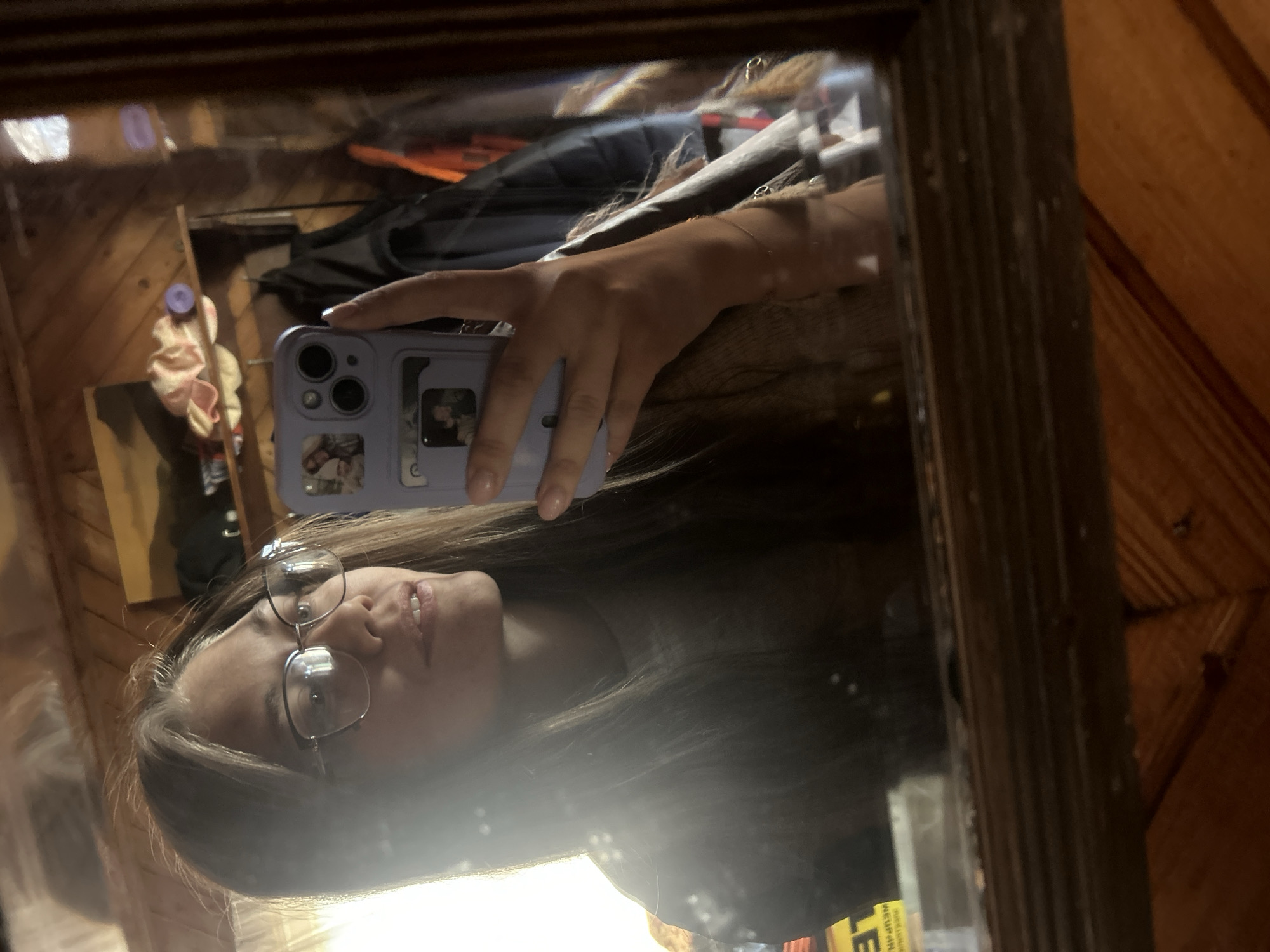 A young person is taking a selfie in a mirror. The person is wearing glasses and holding a purple phone. The reflection in the mirror shows a cluttered room with wooden walls. There is a hair scrunchie and a book or magazine also in the mirror's reflection. The location is undeterminable based on the image.