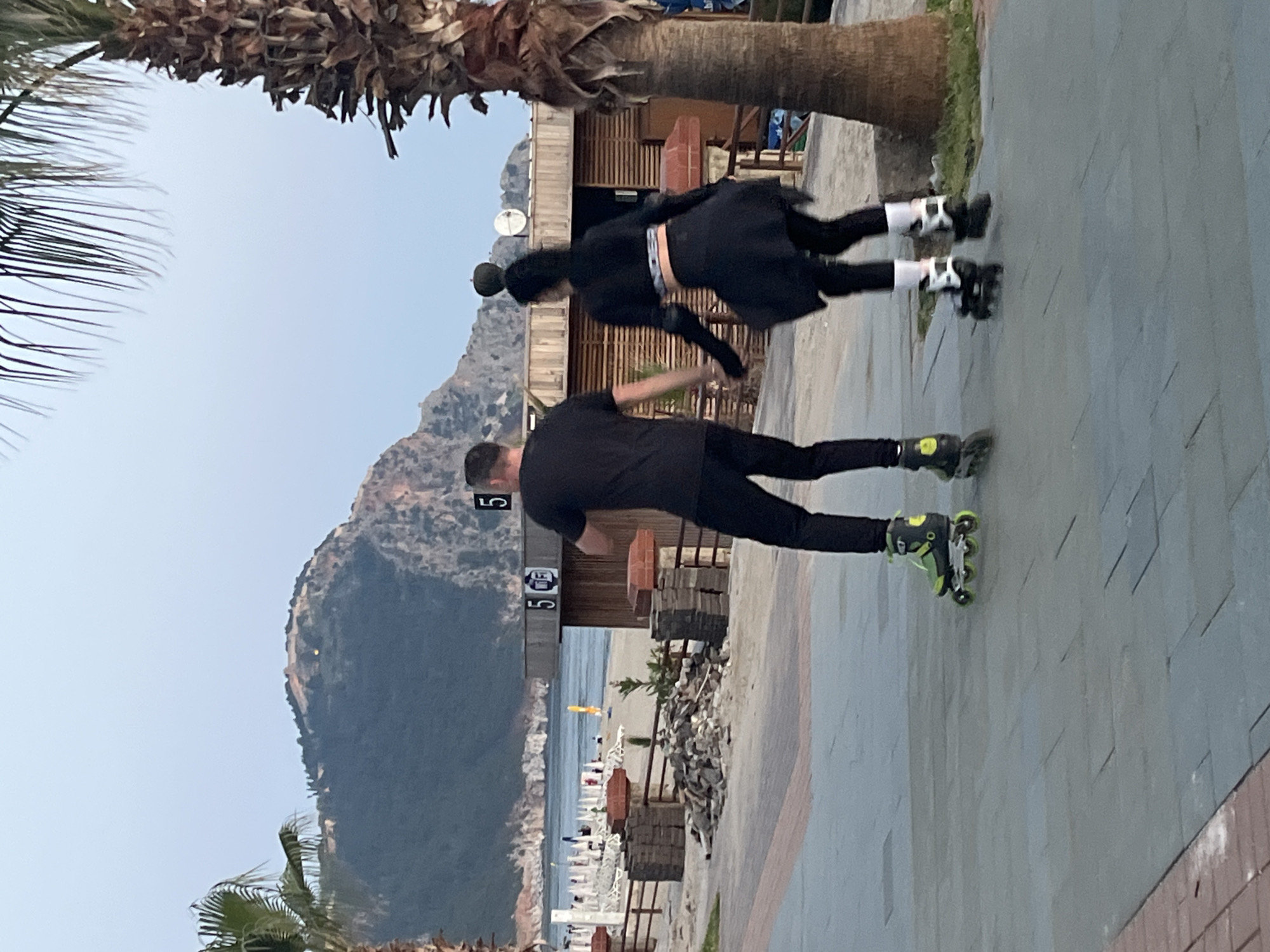 Two individuals, a man and a woman, are captured on a paved outdoor pathway, both wearing inline roller skates. The image is taken from behind them. The man, in the foreground, wears a black t-shirt and black trousers, with distinctive black and lime-green inline skates. The woman behind him wears a black long-sleeved top, a black skirt over black leggings, and black and white inline skates. She also appears to be wearing a black hat or has her hair in a bun.
The scene is set alongside a beach, visible to the right of the subjects, with rows of white sun loungers and umbrellas extending towards the blue sea. Wooden structures, possibly cafes or changing rooms, line the beach side of the pathway. Two white signs attached to these structures clearly display the numbers "5" and "E5". In the distant background, majestic mountains rise under a clear, light blue sky. Palm trees are visible in the foreground and background, framing the view. The ground is made of grey and beige paving stones. The lighting suggests it is daytime, possibly late afternoon or early evening.