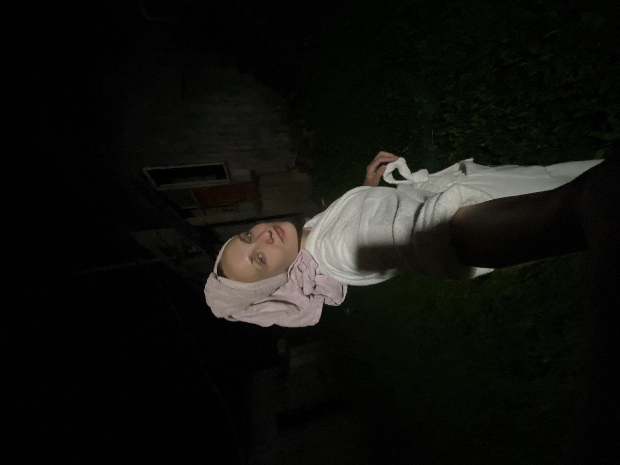 The image shows a woman with a pink towel wrapped around her head and a white bathrobe. She is standing outside at night, with a dark background that appears to be a yard with grass and trees. In the background, there is a building with a window and a door, which is partially illuminated by a light source from the front. The woman is looking towards the camera and smiling. The overall scene is dimly lit, with the exception of the woman and the building. The location is specified as Sharkan, Russia.