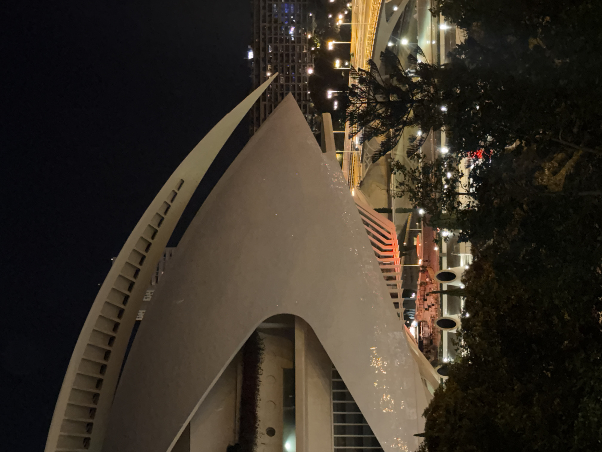 The image shows a nighttime exterior view of the Hemisfèric, a landmark building located in the City of Arts and Sciences in Valencia, Spain. The building, designed by Santiago Calatrava, is characterized by its white, curved structure that resembles an eye or a ship. The foreground is dominated by the imposing white architectural element, illuminated from within and reflecting light. Below and behind the main structure, the urban landscape of Valencia is visible at night. Multiple streetlights illuminate roads and bridges, and the lights of apartment buildings create a textured background. Trees in the foreground partially obscure the view of the city. The overall impression is one of modern architecture set against a backdrop of a vibrant, illuminated city at night.