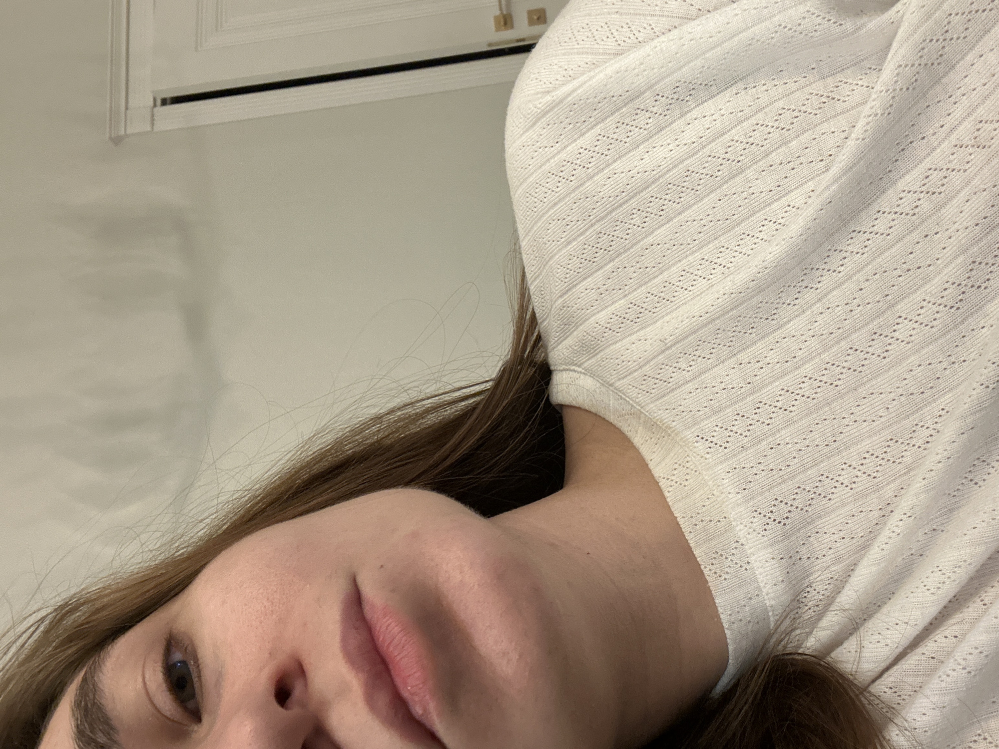The image depicts a close-up, angled view of a single individual, identified as female. Her right eye, eyebrow, nose, and upper lip are visible in the upper left portion of the frame, alongside her chin and jawline on the lower left. Her hair, light brown in color, frames her face and extends over her right shoulder. She is wearing a light-colored, likely white, long-sleeved top featuring a textured, open-knit pattern of diagonal lines and small diamond-shaped perforations. The background consists of a plain, light-colored wall. To the right, a segment of a white door with a molding trim and a metallic, possibly brass-colored, handle is visible. The door appears slightly ajar, revealing a dark vertical gap. The specific location within the city of Kharnavy, Russia, cannot be determined from the visual information provided. No discernible actions or interactions are taking place.