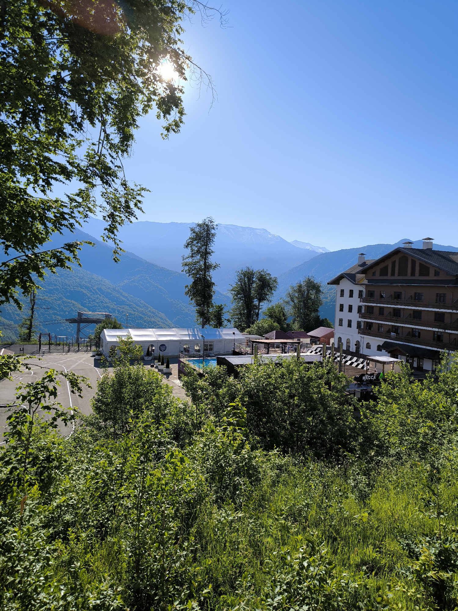 The image depicts a scenic view of a resort area in Estosadok, Russia, nestled within lush green mountains under a clear blue sky. In the foreground, dense foliage of trees and grass frames the scene. Beyond the greenery, a paved area, possibly a parking lot, is visible. Several buildings and structures are situated on a slope, including a large white tent, a swimming pool, and a multi-story hotel with a brown and white facade. In the distance, majestic mountains form a layered backdrop, with some peaks appearing snow-capped. The sun is shining brightly, casting a lens flare in the upper left corner of the image. The overall atmosphere is tranquil and inviting, suggesting a pleasant day at a mountain resort.