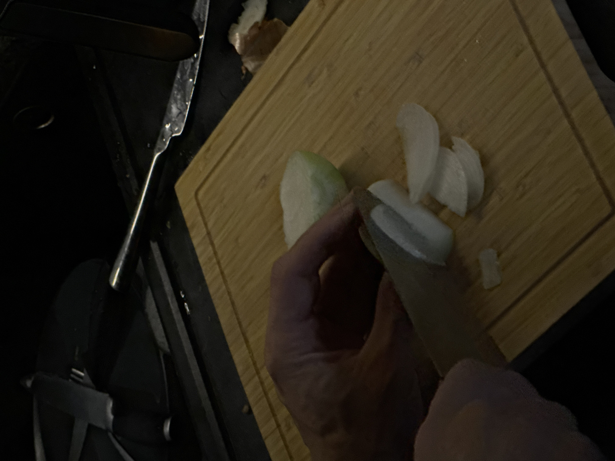 A close-up, dimly lit shot shows a person's hands chopping an onion on a wooden cutting board. The onion is partially sliced, with several thin pieces already cut. A knife is in the process of making another cut. The background is dark and indistinct, with some kitchen utensils visible. There is no identifiable information to place this scene in Bobrovo, Russia.
