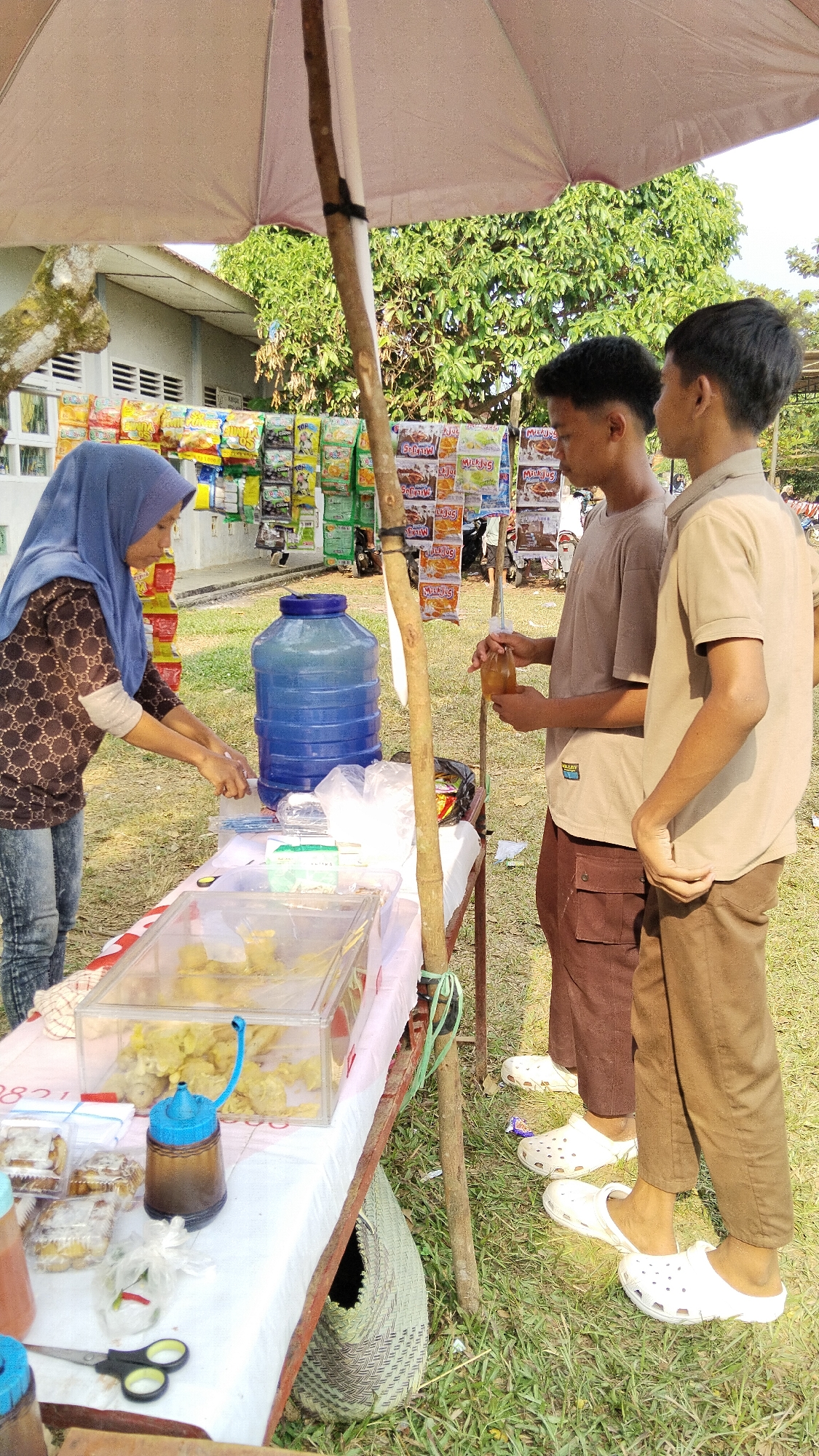 An outdoor scene in Margatunggal, Indonesia, captures a vendor selling snacks and drinks under a large, light-colored umbrella. The vendor, a woman wearing a blue hijab and a patterned brown and white long-sleeved shirt, is interacting with two young males. She is leaning over a table, seemingly attending to a transaction or preparing an order. One young male, wearing a brown t-shirt and brown cargo pants, holds a small bottle with a brown liquid, possibly a drink or sauce. The other young male, wearing a tan t-shirt and brown pants, stands beside him. The table is covered with a white cloth and displays various items including a clear plastic container filled with fried snacks, a large blue water dispenser, plastic-wrapped food items, and a pair of scissors. A woven basket is placed on the ground beneath the table. The background shows a building with a white facade and rows of packaged snacks displayed on racks. Trees with green foliage are visible behind the stall, and the ground is covered in grass. The overall impression is that of a casual, local market or roadside stall.