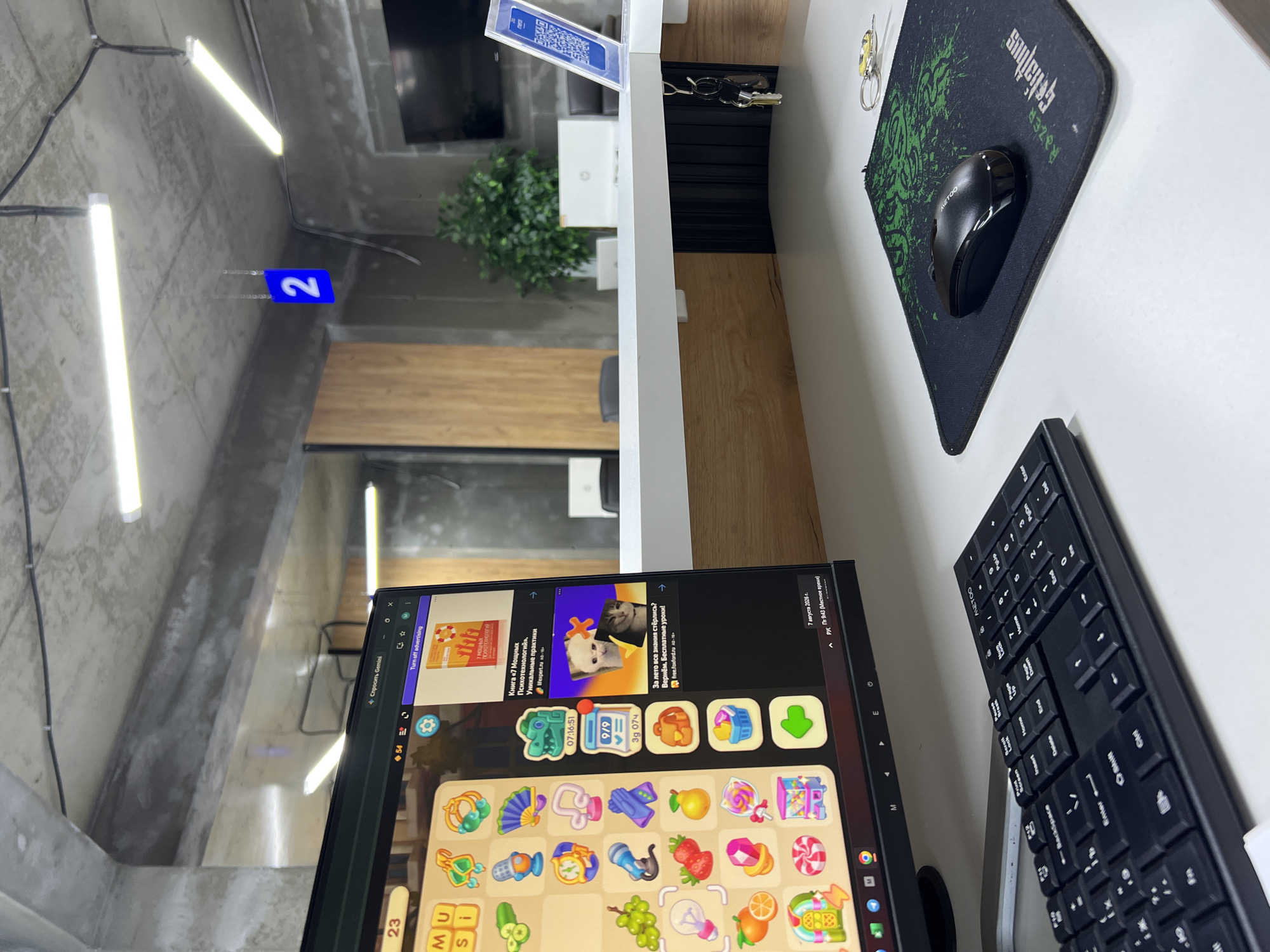 The image depicts an indoor scene, likely an office or co-working space, with a focus on a desk setup. In the foreground, a black keyboard and a black mouse on a green and black patterned mousepad are visible. To the left, a computer monitor displays a colorful game interface with various icons resembling game pieces and cartoon characters. There are also advertisements visible on the right side of the monitor screen, one promoting a book about psychology and another featuring cats.
The desk itself is white and appears to be part of a larger counter or workstation. Below the desk surface, wooden paneling and black ribbed material are visible. Keys are hanging from a hook on the underside of the desk.
In the background, the environment is industrial in style with exposed concrete ceilings and fluorescent lighting. A large potted plant is situated behind the desk, and a television screen is mounted on the wall. A blue sign with the number "2" is hanging from the ceiling.
The image does not contain any explicit details that would indicate its location in the city of Shahrixon, Uzbekistan.