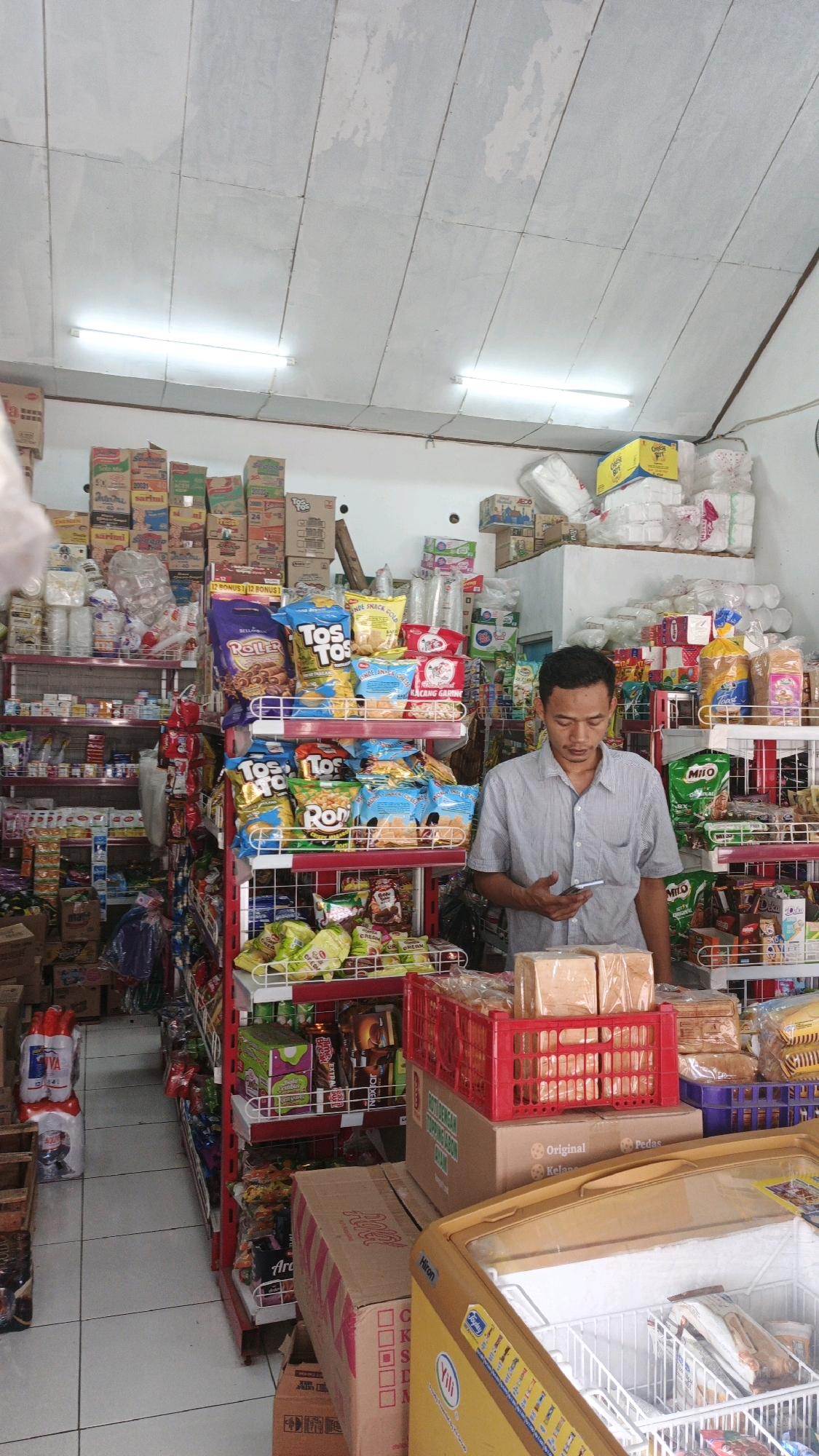 A man stands in a small convenience store in Cibenda, Indonesia, holding a smartphone and looking down at it. He is wearing a light blue and white striped short-sleeved shirt. The store is filled with shelves stocked with various grocery items, including snacks, drinks, and packaged food. In the foreground, there are stacks of bread in red and blue crates, a cardboard box, and the top of a yellow freezer. The ceiling is made of white panels with fluorescent lights. The floor is tiled.
