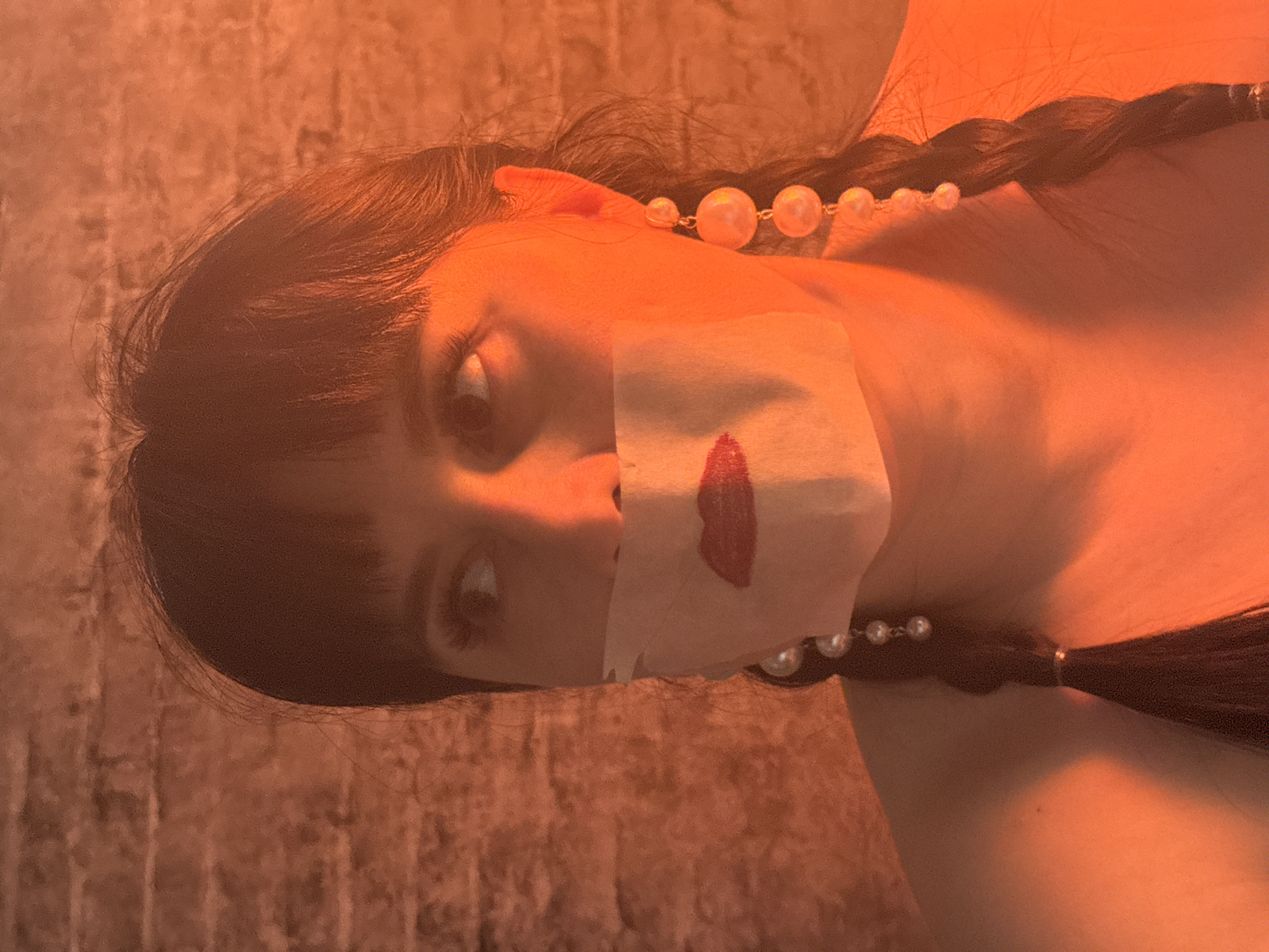The image, a portrait photograph rotated 90 degrees counter-clockwise, features a woman bathed in an intense reddish-orange light. Her dark hair is styled in braids, one of which is adorned with a string of pearls that extends from near her ear. A piece of light-colored material, resembling paper or tape, is affixed across her mouth, bearing a prominent smear of red lipstick or paint, suggesting a silenced or muffled voice. Her wide, dark eyes gaze directly at the viewer, conveying an intense or perhaps unsettling emotion. The background is a textured surface, possibly a brick or stone wall, completely saturated in the same warm, orange hue, contributing to an overall moody and surreal atmosphere. There is no visible text. While the location context is Zav'yalovo, Russia, no specific visual cues within the image identify this location. The dominant warm lighting makes the time of day and outdoor weather conditions impossible to determine.