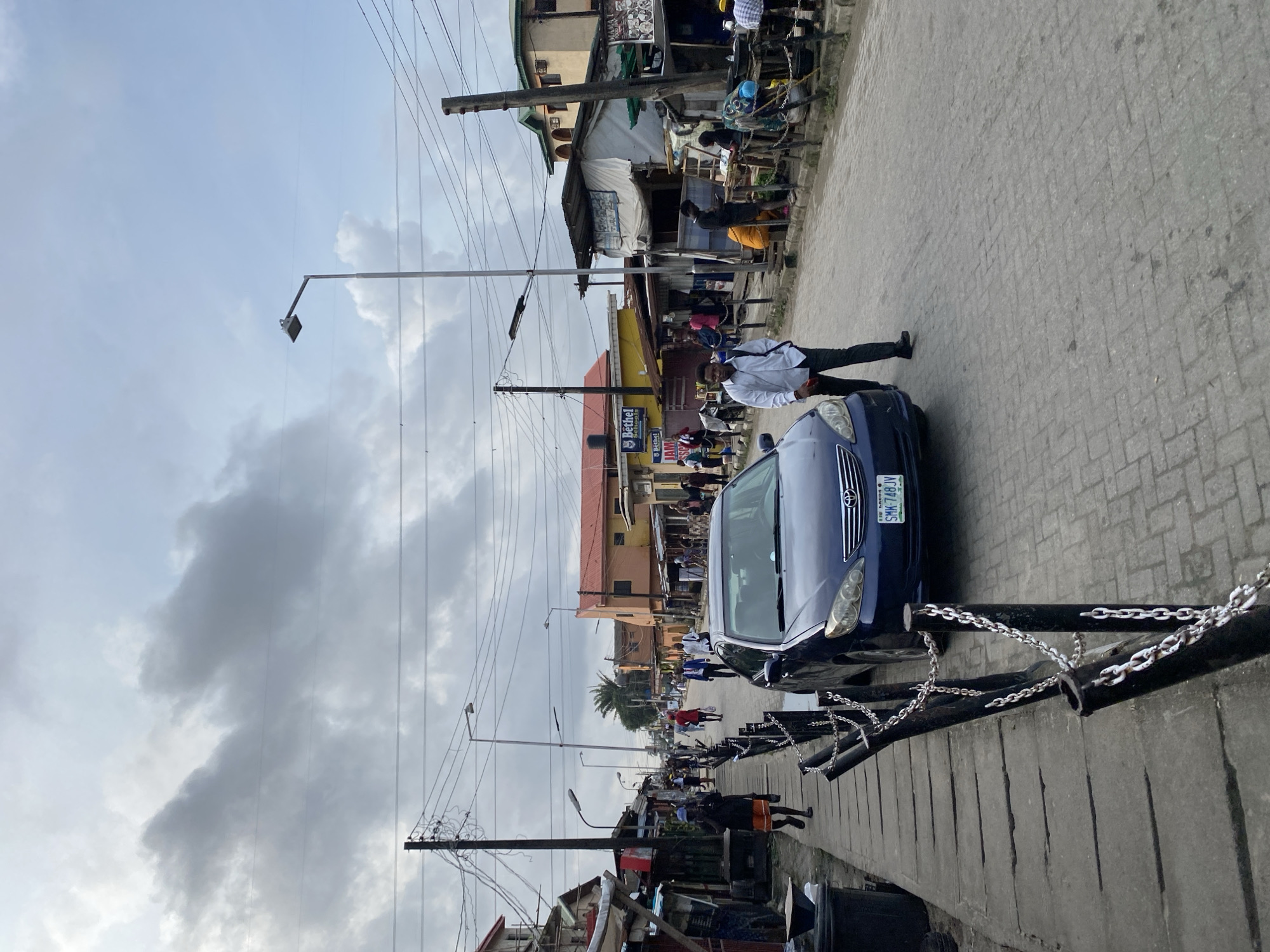 The image shows a street scene in a city. A blue sedan is parked on the right side of the road, which is paved with interlocking bricks. A man in a white shirt and dark pants is standing next to the car. Several other people are walking on the sidewalk to the left of the road, which is made of concrete slabs. There are buildings on both sides of the street, some of which appear to be shops or market stalls. Overhead, there are power lines and streetlights. The sky is overcast with gray clouds. The scene appears to be a busy urban environment with pedestrian and vehicular traffic.