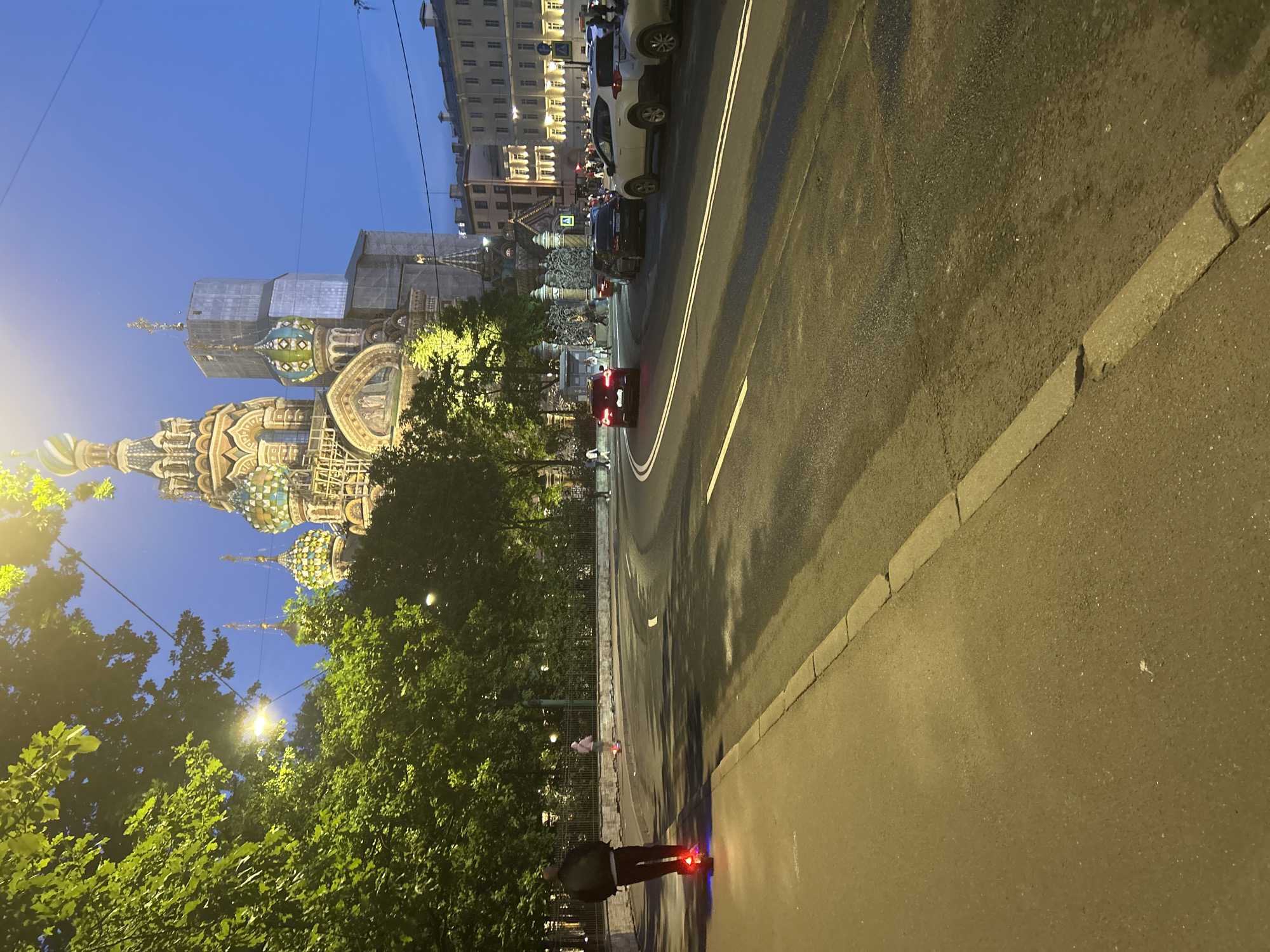 A person is riding an electric scooter on a sidewalk at night. The scooter has red taillights. The sidewalk is made of asphalt and bordered by a concrete curb. A street with cars and streetlights is to the right of the sidewalk. In the background, a large, ornate church with colorful domes and a cross-topped spire is illuminated. The sky is dark blue, indicating evening or night. Trees line the left side of the sidewalk.