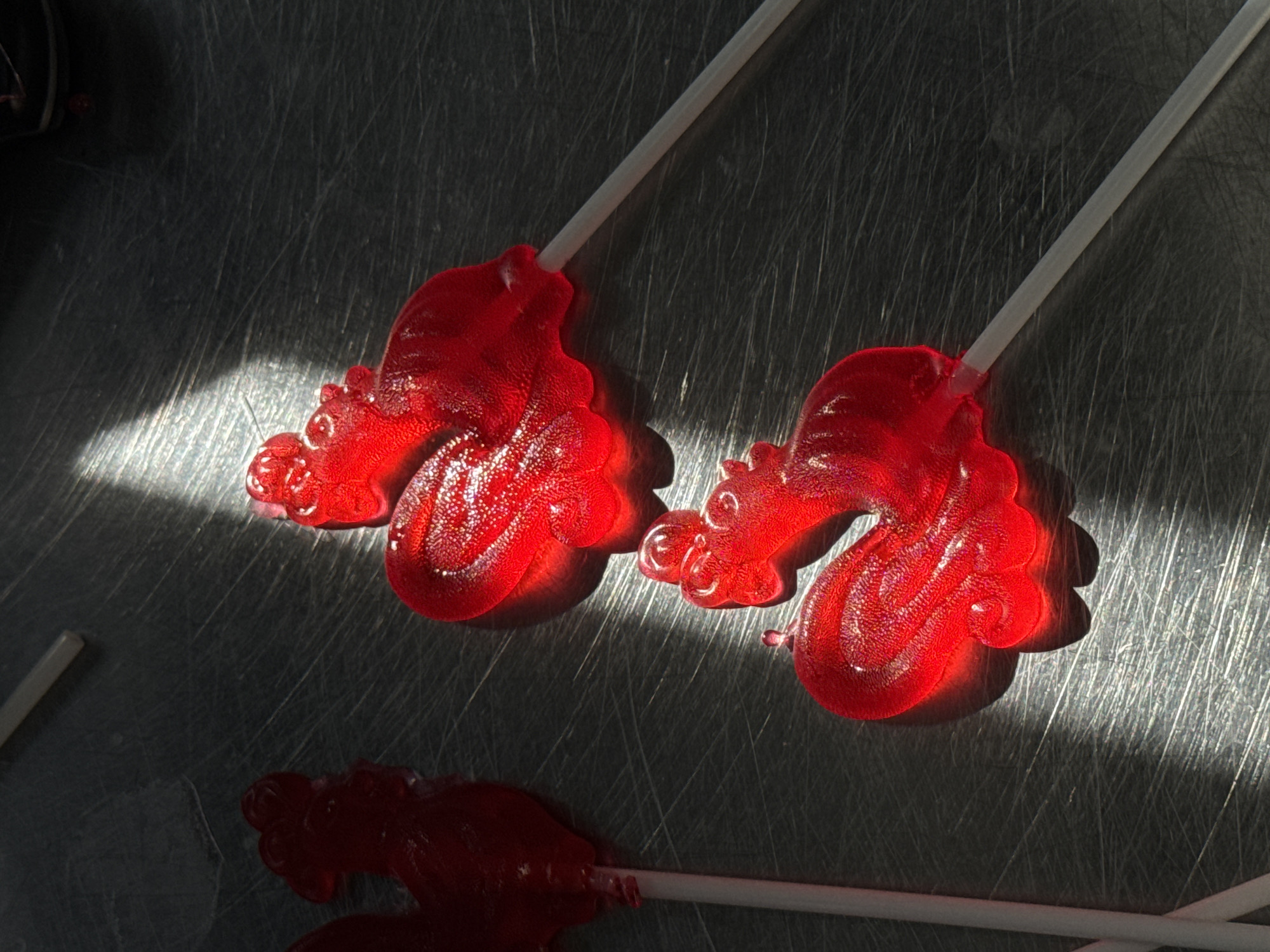 The image shows three red rooster-shaped lollipops on a metallic surface with visible scratches. The lollipops have white sticks. A strong light source illuminates the top two lollipops, casting distinct shadows. The bottom lollipop is partially in shadow. There are no people present. This image does not include any data that can place it in Perm, Russia.
