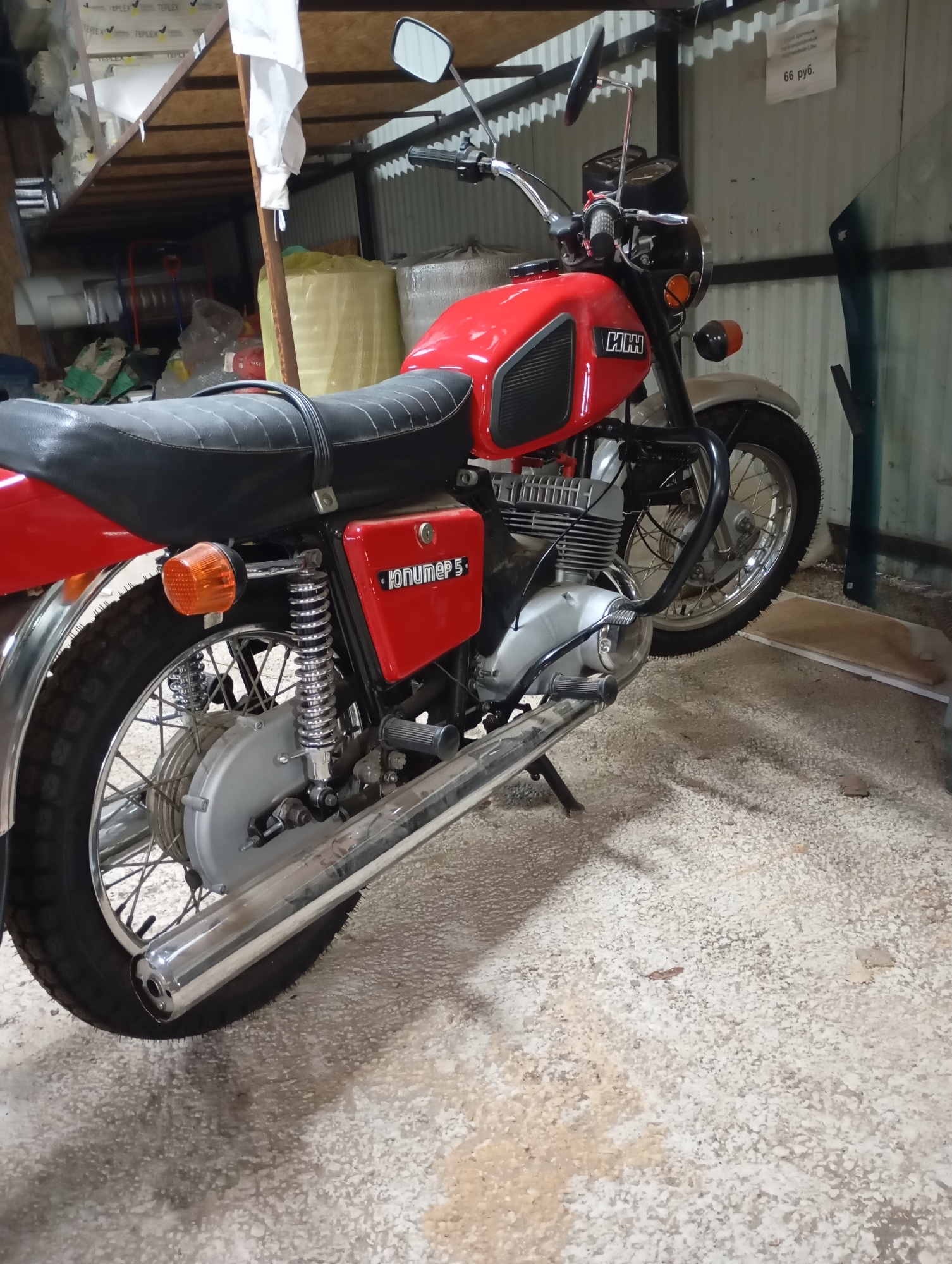 The image displays an indoor scene within a storage or workshop facility in Фоки, Russia. A red IZh Jupiter 5 motorcycle is the central subject, positioned with its side stand deployed, facing towards the right and slightly away from the viewer. The motorcycle features a red fuel tank with black rubber knee pads and "ИЖ" (IZh) branding, a black ribbed seat with a black securing strap, and chrome components including the exhaust pipe, spoked wheels, handlebars, and rear shock absorbers. Orange turn signal indicators are visible at both the front and rear.
The surrounding environment indicates an industrial or utilitarian space. In the upper left background, metal and wooden shelving units are stacked with multiple white, rectangular items, some labeled "TEPLEX". Below these shelves and further back, various large bags and wrapped materials, including a prominent yellow roll, are stored. The walls are constructed from light-colored corrugated metal sheeting, punctuated by dark structural beams. On the right side of the frame, a large pane of green-tinted glass, possibly a vehicle windshield, rests against the wall. A white paper sign, attached to the wall, displays the text "66 руб." (66 rubles). The floor is a light-colored concrete surface, heavily textured with a covering of small, white, granular debris or dust. No individuals are present in the scene.
