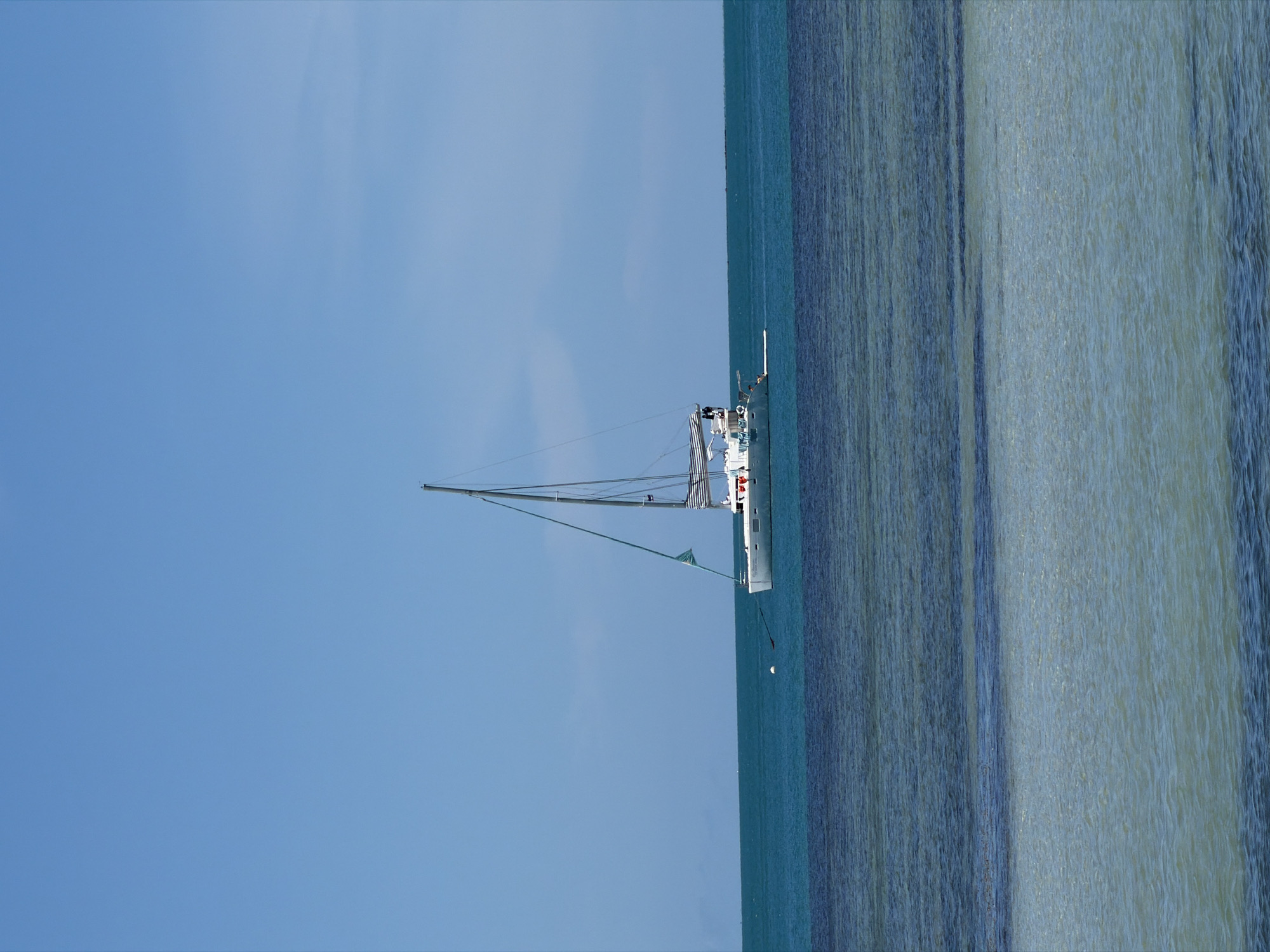 A white sailboat with a tall mast and a partially unfurled sail is depicted at sea under a clear blue sky. The sailboat is positioned near the right edge of the frame, with its hull visible against the turquoise water. The water exhibits a striped pattern, suggesting ripples or currents. The image is oriented vertically, with the sky occupying the left half and the sea the right half. There are no visible people, text, or specific details to indicate the time of day or weather conditions beyond the clear sky. The location context provided suggests this scene is in Baan Thong Krut, Thailand.