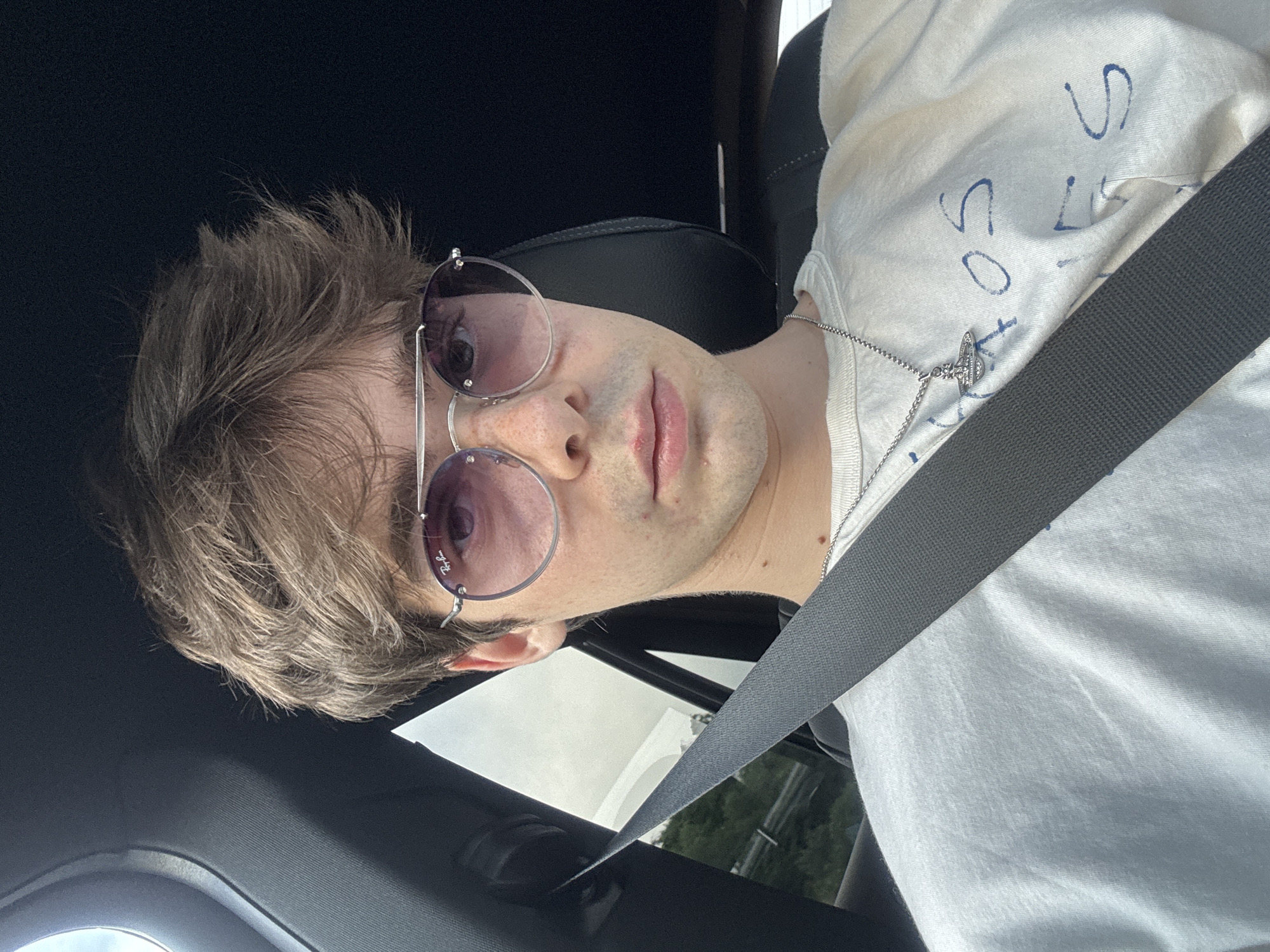 A young Caucasian male with light brown, tousled hair and freckles across his nose and cheeks is pictured from the chest up, looking directly at the camera. He is wearing large, round, silver-rimmed sunglasses with pink-tinted lenses and a white t-shirt with blue writing. A silver chain with a Saturn pendant is visible around his neck. He is seated in the driver's seat of a vehicle, with a grey seatbelt diagonally across his chest. The interior of the car is visible, with dark upholstery and a glimpse of greenery and a cloudy sky through the window on the left side. The scene appears to be set on a road, possibly on the outskirts of Bela Cerkev, Slovenia, given the visible vegetation and highway infrastructure in the background.