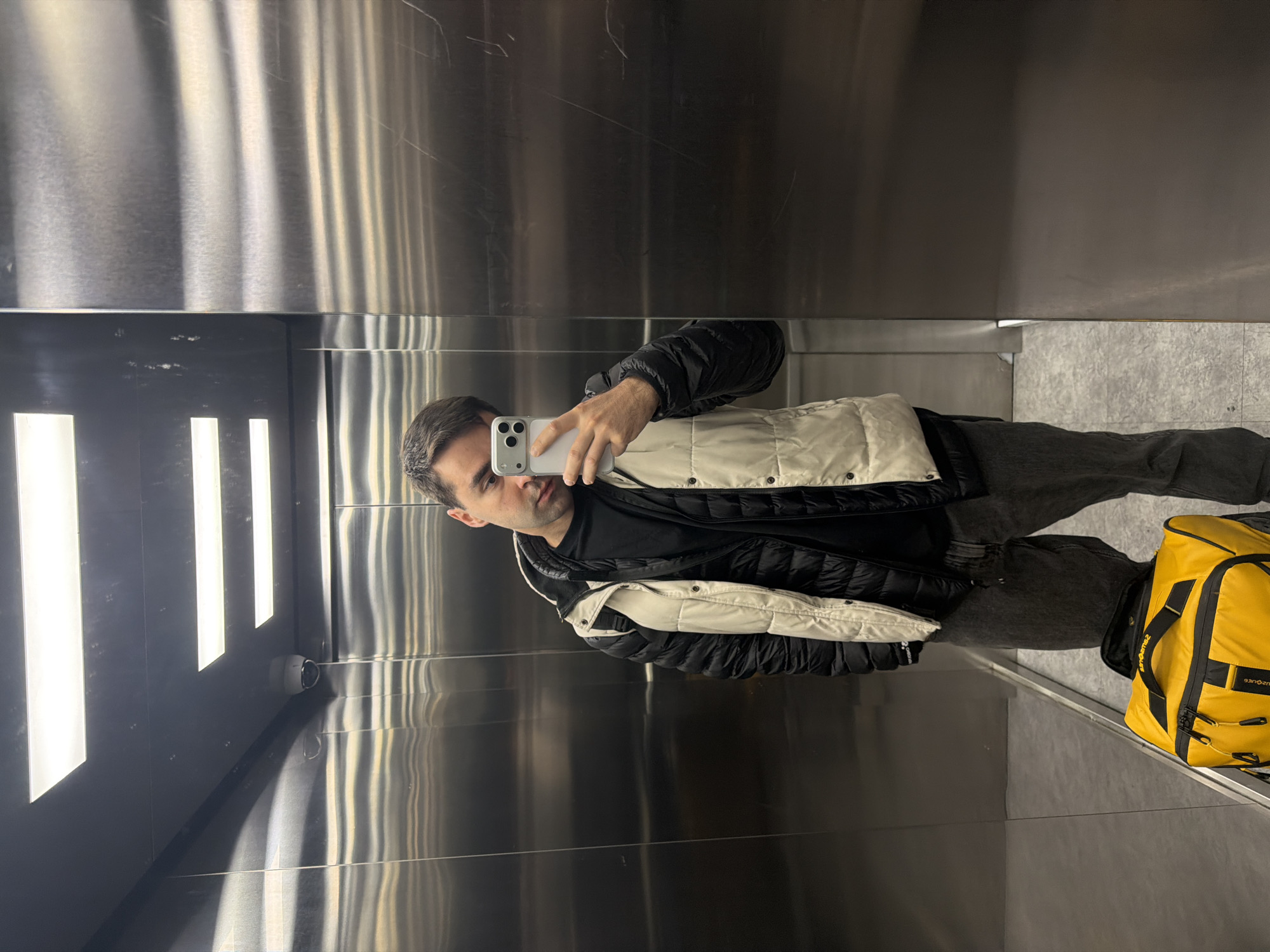 A person is taking a mirror selfie inside an elevator. The person is wearing a black t-shirt, grey jeans, and a puffy jacket that is primarily black with cream-colored sections. They are holding a white smartphone in their left hand, and their face is visible in the reflection. Next to the person is a large yellow duffel bag with black straps.
The elevator interior consists of brushed metal walls with vertical grain. The left side of the elevator has illuminated vertical light bars. A security camera is mounted on the wall, below the light bars. The floor appears to be a neutral gray color.
There is no information in the image to indicate this photograph was taken in Tbilisi, Georgia.