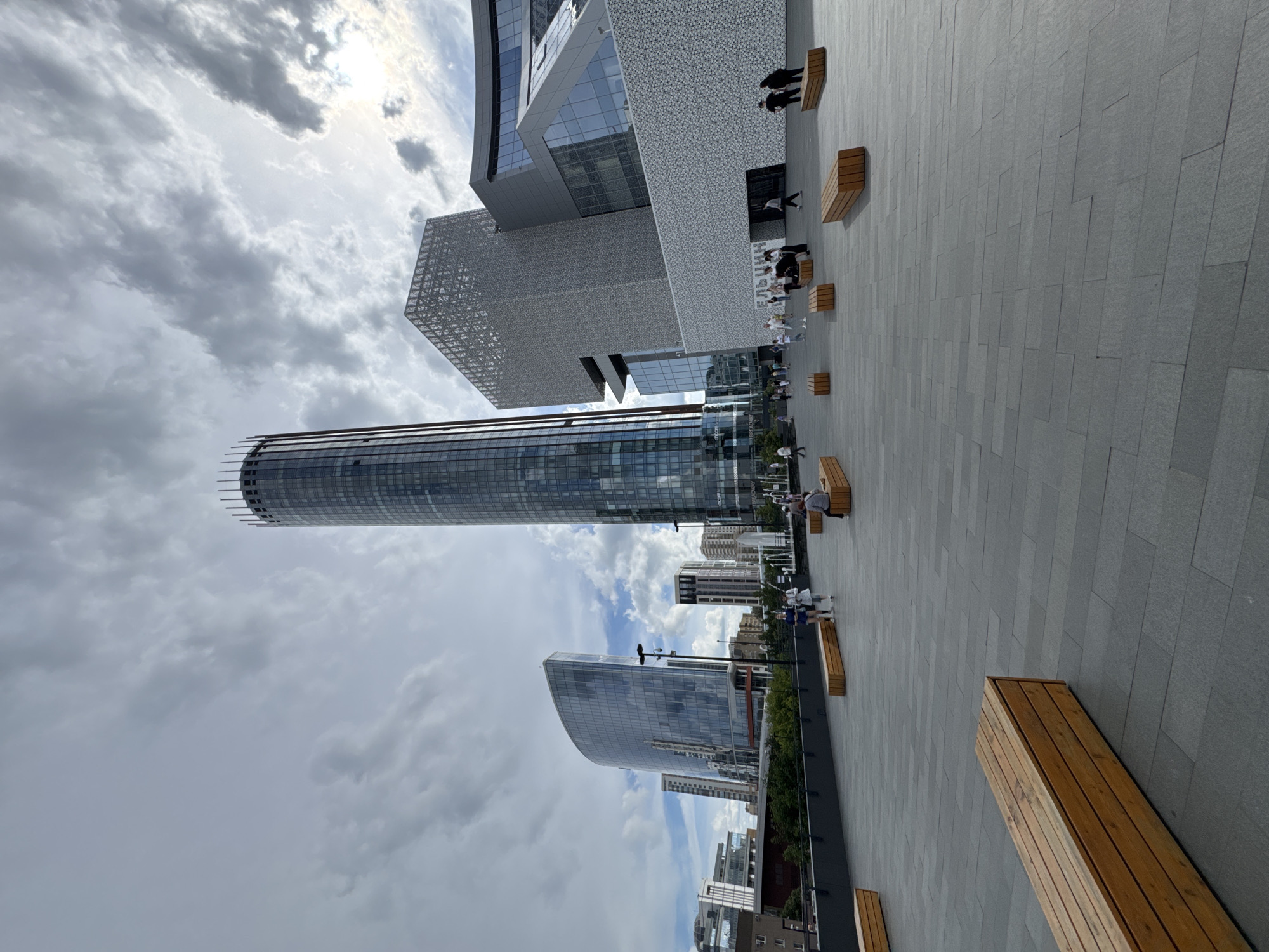 This media file captures an outdoor scene in Yekaterinburg, Russia, under a cloudy sky with some areas of bright sunlight peeking through. The perspective is from a paved plaza with a patterned grey stone floor. Several modern buildings dominate the background.
Prominently featured is a tall, cylindrical skyscraper with a reflective glass facade. Adjacent to it, a more angular building with a patterned metallic exterior is visible. Further in the distance, other contemporary high-rise structures add to the urban landscape. The scene appears to be an urban development or business district.
Several wooden benches are placed on the plaza, with a few people seated and standing on or near them. A small group of individuals is walking on the plaza, suggesting a casual daytime activity. The overall mood seems relaxed and ordinary. No specific event is discernible, and the time of day appears to be daytime, given the lighting. The weather is mixed, with clouds and sunshine.