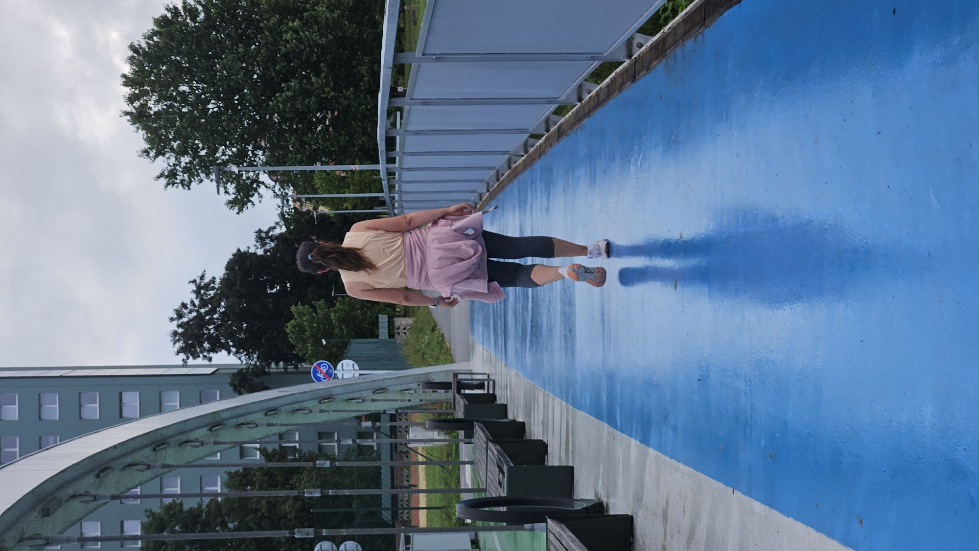 The image shows a woman walking away from the camera on a wet, bright blue elevated pedestrian and cyclist path. She is wearing a light peach-colored tank top, dark grey capri leggings, and white athletic shoes with orange accents. A pink hooded jacket is tied around her waist. The path is lined with a light blue metal railing on the right and has wooden benches on the left. In the background, a modern, multi-story building with a curved facade and visible windows is present, along with trees and a cloudy sky. This scene is located on the bridge over the Sekčovský stream in Prešov, Slovakia, specifically the part of the bridge accessible from the park near the residential area of Sídlisko III.