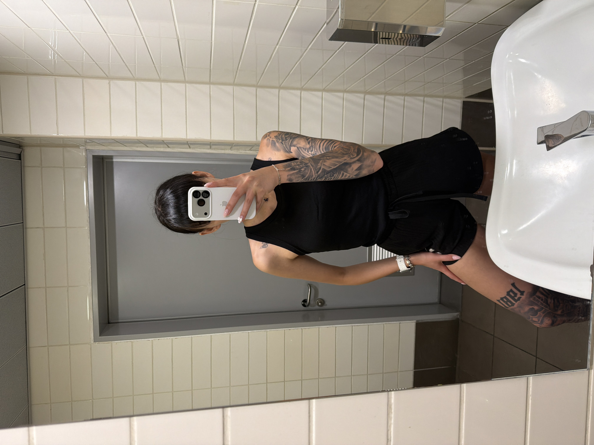 An individual is captured in a mirror selfie inside what appears to be a public restroom. The specific location within Plovdiv, Bulgaria, cannot be determined from the visual evidence.
The subject is a person, likely female, with dark hair pulled back, featuring a fringe/bangs. The face is obscured by a white iPhone, held in the left hand, displaying a triple camera system and the Apple logo. The individual is wearing a black ribbed tank top and black drawstring shorts. A white watch is visible on the left wrist, and a silver chain bracelet is on the right wrist. A small, dark tattoo resembling a butterfly or insect is present on the left upper shoulder/arm area. A full sleeve tattoo, dark and intricately patterned, covers the right arm. A prominent, dark, old English style tattoo, possibly spelling "angel" or a similar word, with additional design elements, is visible on the left upper thigh/knee.
The immediate environment consists of cream-colored rectangular wall tiles with dark grout lines, extending from the floor to the ceiling. A grey door with a chrome handle is visible in the reflection behind the subject. The floor is covered with dark brown/grey tiles. A white ceramic sink with a chrome faucet is in the foreground. To the right, a silver wall-mounted dispenser (likely for soap or paper towels) is affixed to the tiled wall. The ceiling features grey perforated acoustic panels.