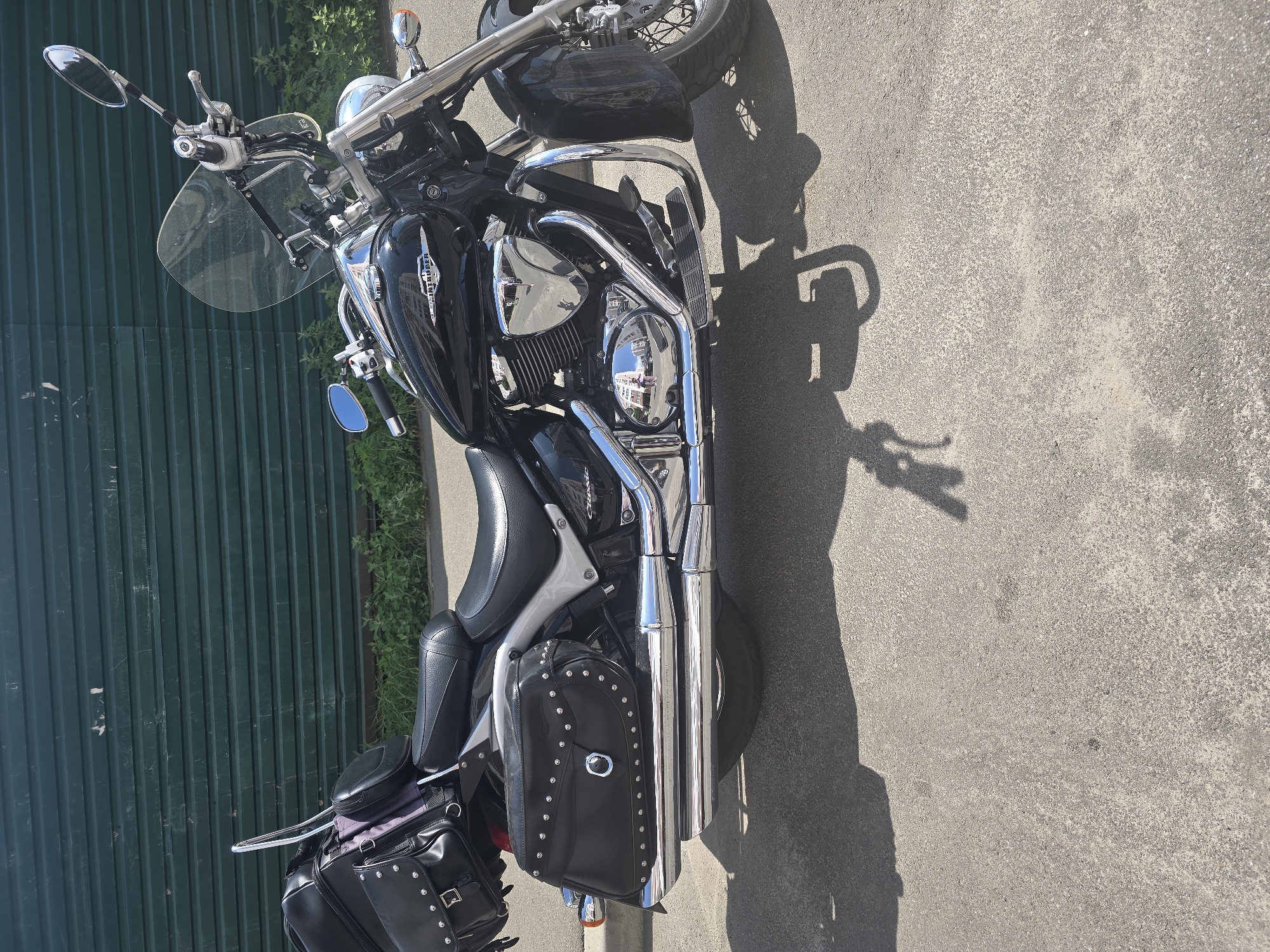 In Vyshneve, Ukraine, a black Suzuki Intruder motorcycle is parked on an asphalt surface. The motorcycle features black leather saddlebags adorned with silver studs, a passenger backrest with a black luggage bag, and a clear windscreen. The motorcycle is positioned in front of a green corrugated metal fence. Sunlight casts a distinct shadow of the motorcycle and its rider onto the ground. The scene appears to be outdoors, with a small amount of green vegetation visible between the motorcycle and the fence.