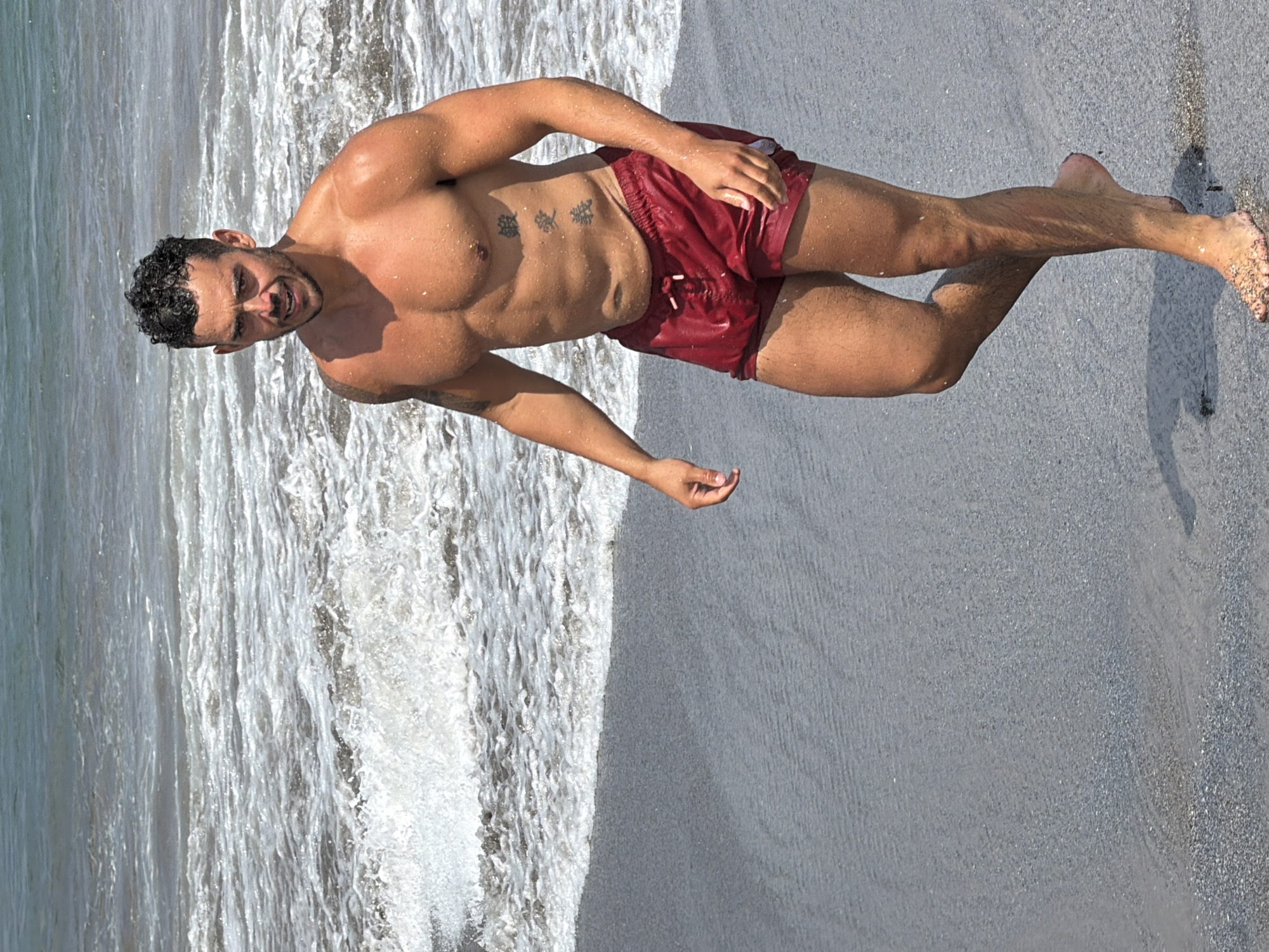 A muscular man with dark, curly hair and a goatee walks along the wet sand of a beach in North Kuta, Indonesia. He is shirtless, revealing tattoos on his chest and arm, and is wearing red swim trunks. The ocean waves are breaking behind him, and the water is a mix of white foam and darker blue. The sand is dark gray and appears wet.