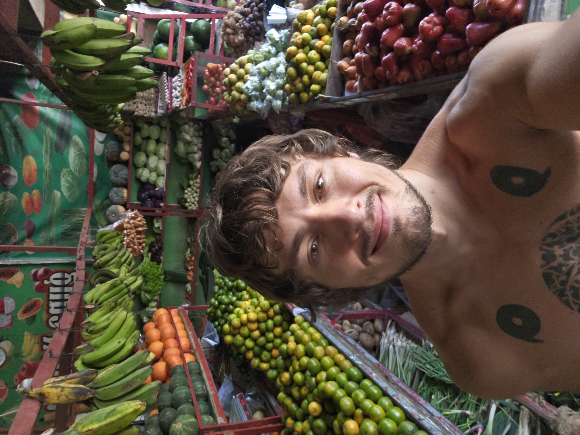 The image is a selfie taken by a light-skinned male with short, curly brown hair and a trimmed beard. He is topless, revealing a tattoo on his upper left chest and another on his upper right shoulder. The background is a vibrant fruit stand, filled with various fruits, including plantains, bananas, oranges, rose apples, and small green citrus fruits. The stand is structured with red metal frames and shelves. A green backdrop with printed fruit images is visible behind some of the produce. The setting appears to be a bustling market. The city and country are not specified in the prompt.