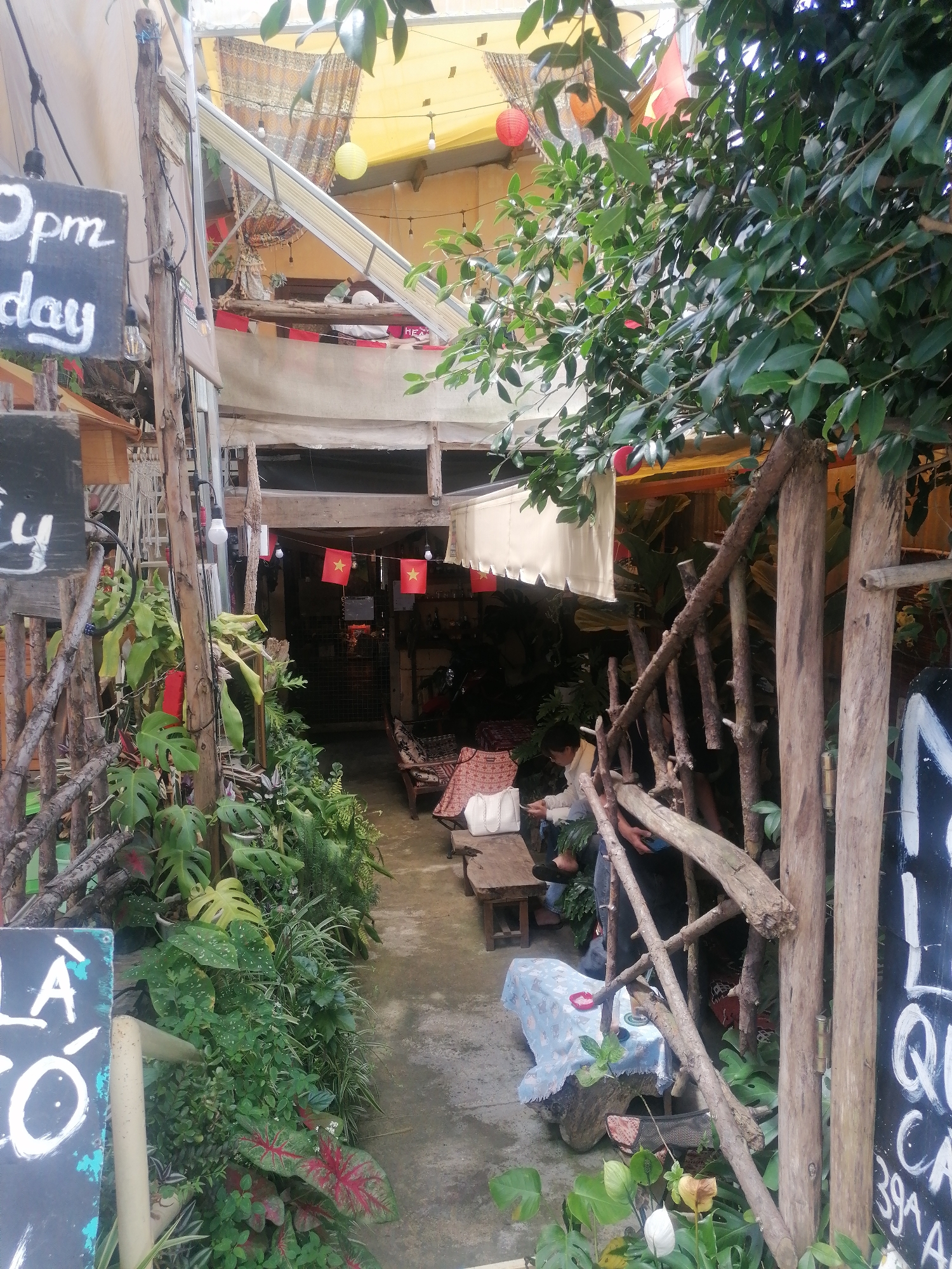 A narrow, outdoor walkway leads into a cafe in Da Lat, Vietnam. The walkway is lined with lush greenery and rustic wooden structures. On the left, a blackboard sign reads "0pm Sunday" in chalk. Further down, Vietnamese flags are strung across an opening. Inside the cafe, people are seated at tables, some interacting with their phones. The atmosphere appears casual and bohemian, with draped fabrics, exposed wood, and natural elements contributing to the cafe's charm.