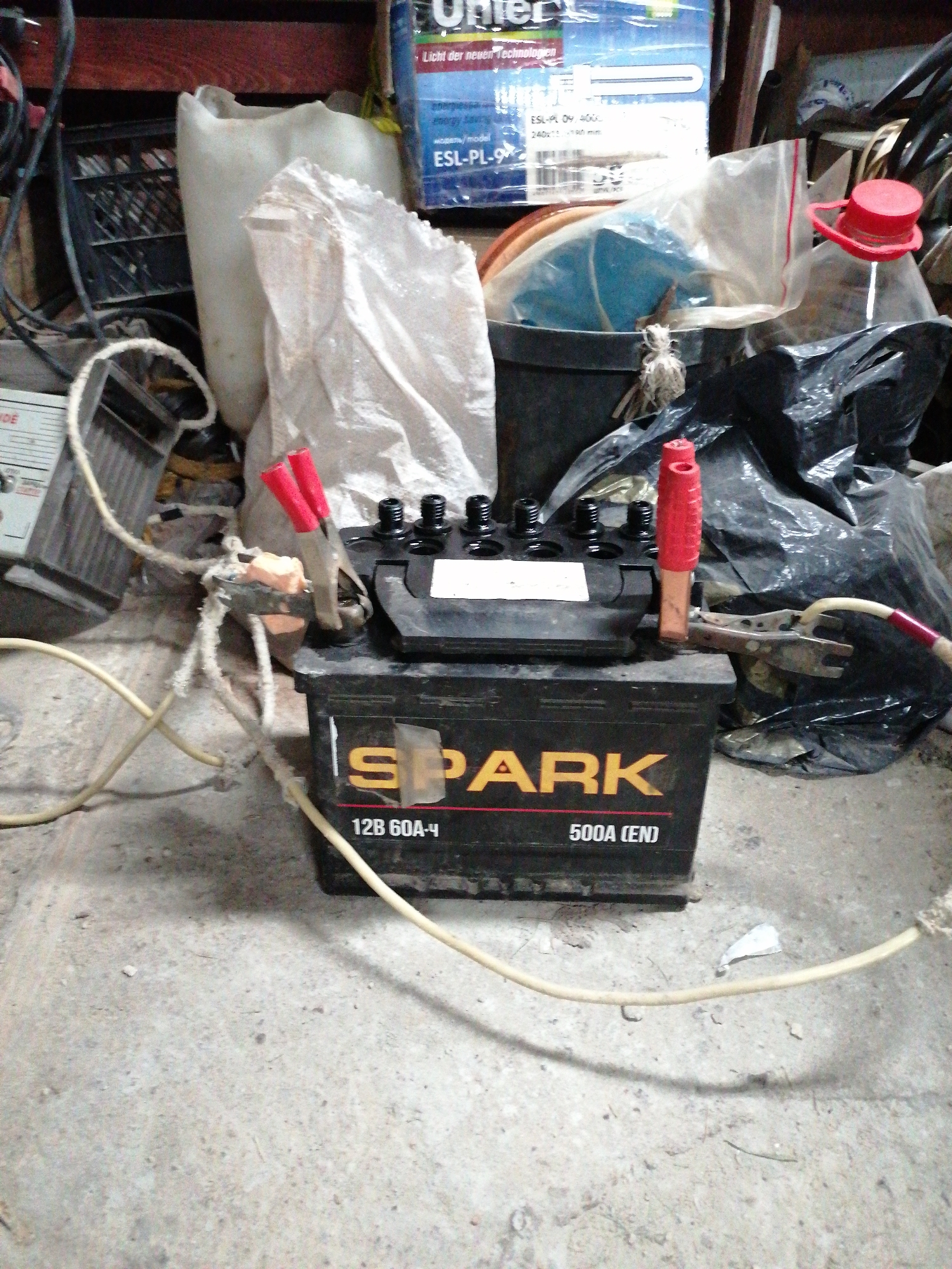 The image shows a black SPARK 12V car battery sitting on a grey concrete floor, with a pair of battery clamps attached to its terminals. Red plastic sleeves cover the handles of the clamps. The battery is labelled "12B 60A-ч 500A (EN)". Several wires covered with a dried, foamy substance connect to the clamps. In the background, there is a variety of items including a black plastic crate, a white plastic container, a white cloth sack, a black bucket, a black plastic bag, and a product box labelled "Unter Licht der neuer Technologien ESL-PL-9". The setting appears to be a cluttered garage or storage area.