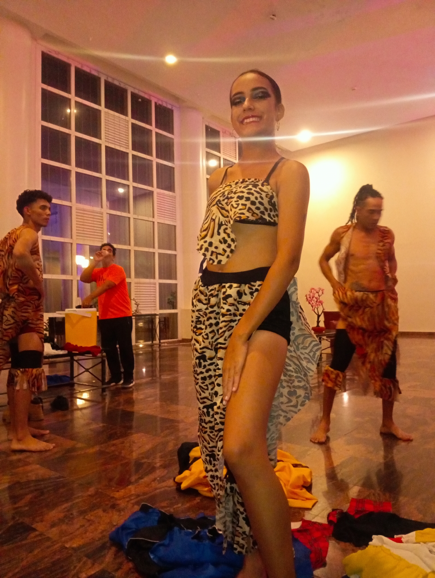 Captured in a room with a shiny marble floor and illuminated by overhead lights, the image features four people. A young woman is prominently positioned in the foreground, wearing a leopard-print outfit and black shorts. To her left stands a man clad in a tiger-striped ensemble, while another man, shirtless but similarly adorned with tiger-striped fabric, is visible to her right. Between the first man and the woman is a man in a red shirt drinking from a glass at a table with a cooler. The floor is scattered with piles of clothing, suggesting a dressing or preparation area. In the background are large windows, hinting at an external environment. Pedro González, Venezuela, is referenced, providing a geographical context for the scene.
