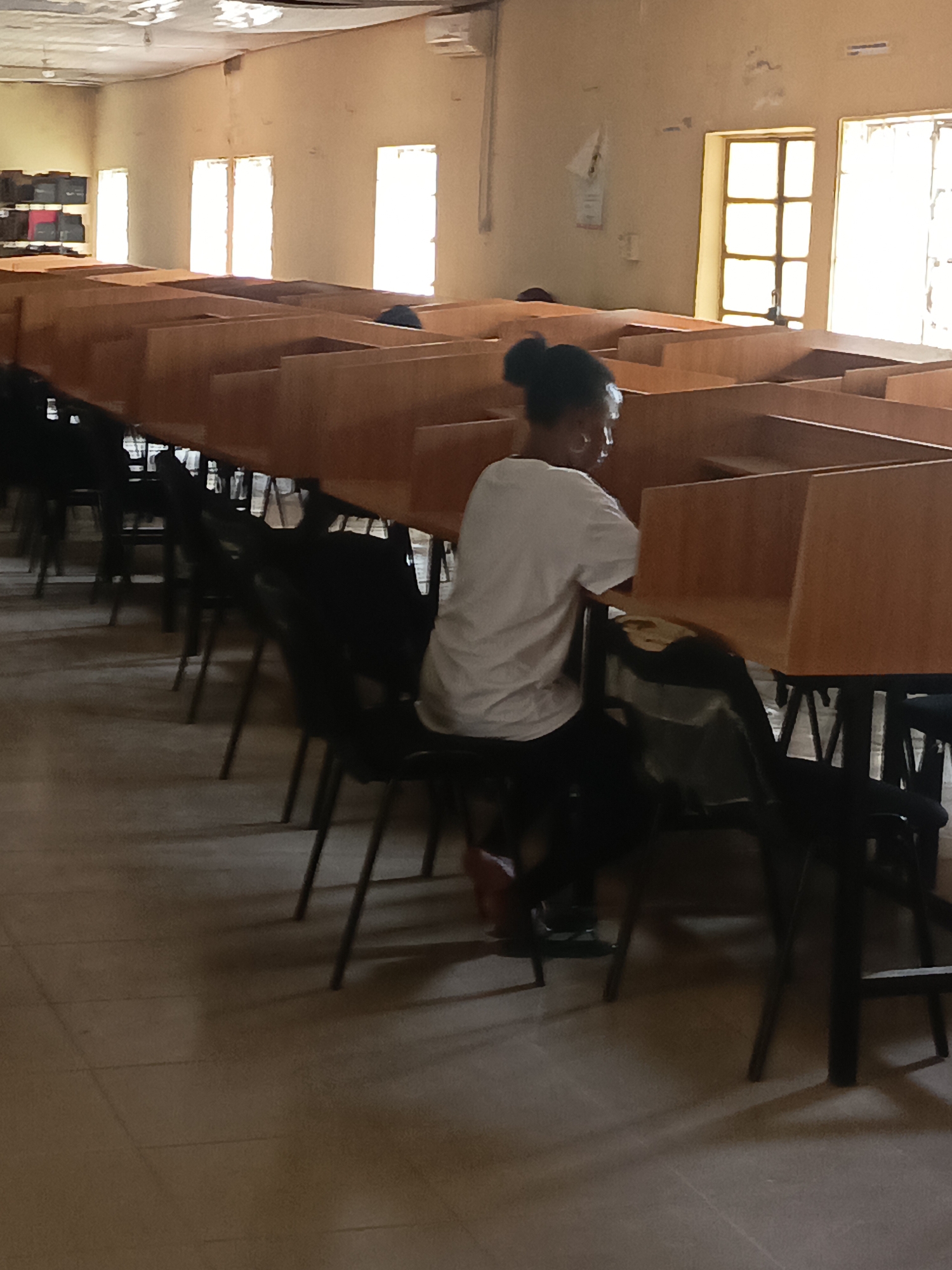 The image depicts the interior of a library in Akwanga, Nigeria. A young woman with her hair in a bun sits at a long row of wooden study carrels. She is wearing a white t-shirt and black pants. The carrels stretch into the distance, with rows of black chairs tucked beneath them. Sunlight streams through multiple windows along the back wall. The library appears to be relatively empty. The floor is tiled. Bookcases with books are visible on the left wall.