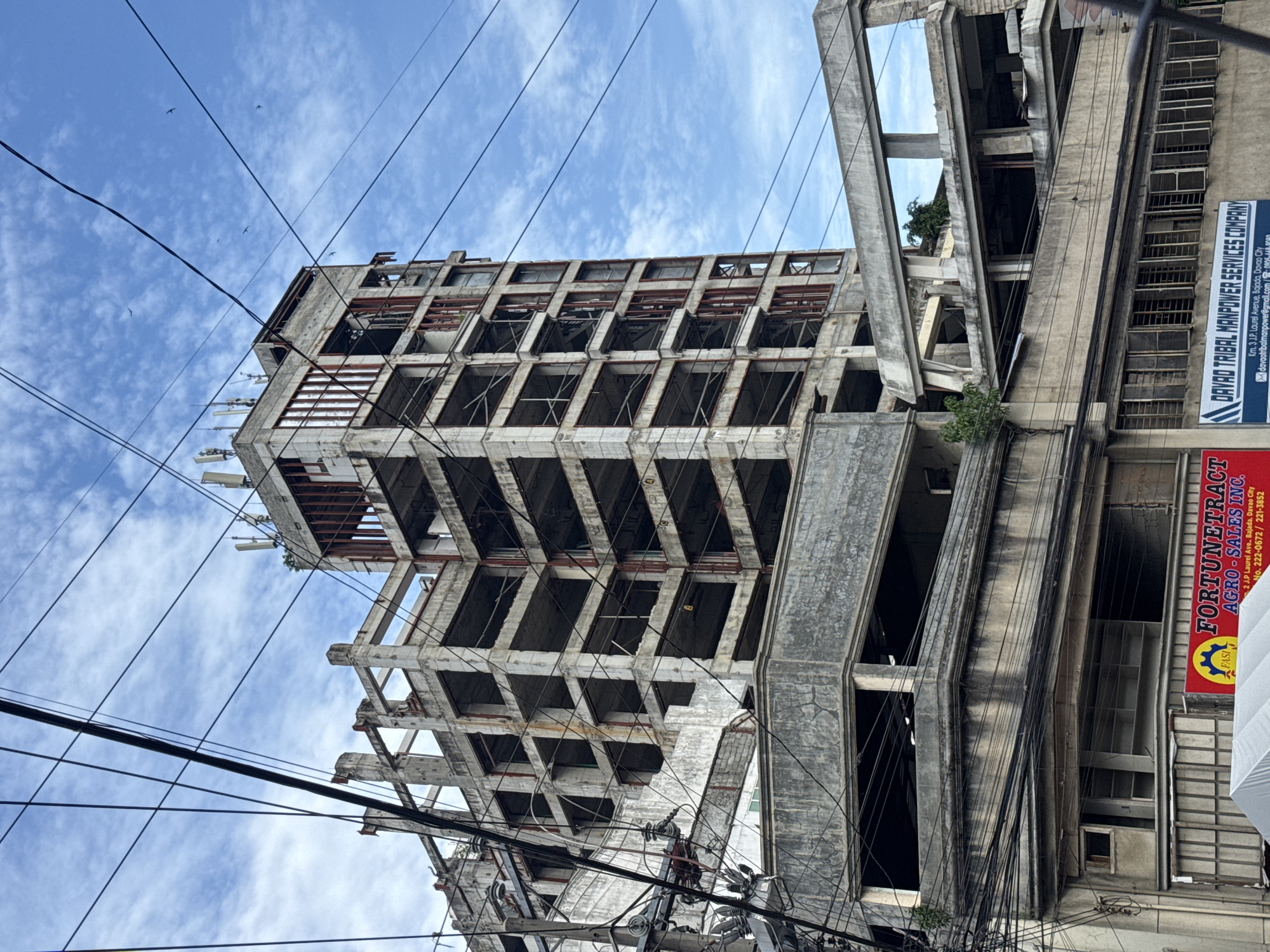 The image depicts an unfinished or dilapidated concrete building in Davao City, Philippines. The building's skeletal structure is exposed, with empty window frames and weathered concrete. Numerous electrical wires crisscross the foreground and partially obscure the building. A business sign for "FORTUNETRACT AGRO-SALES INC." and "DAVAO TRIBAL MANPOWER SERVICES COMPANY" is visible on a neighboring building. The sky above is blue with scattered clouds. The location is identified through the signs, mentioning J.P. Laurel Avenue, Bajada, Davao City.