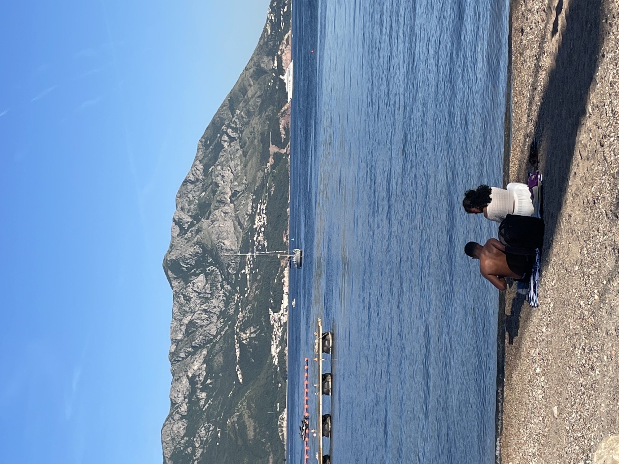 The image shows a view of the Adriatic Sea in Budva, Montenegro, with a mountainous coastline on the left side of the frame. A city with white buildings is nestled along the base of the mountain. In the water, there is a sailboat and a pier-like structure with black objects attached to it. On the right, there is a gravelly shoreline where two individuals are present. One person, wearing a white outfit, is sitting down, while another person, shirtless and wearing dark-colored shorts, is crouched towards the water.
