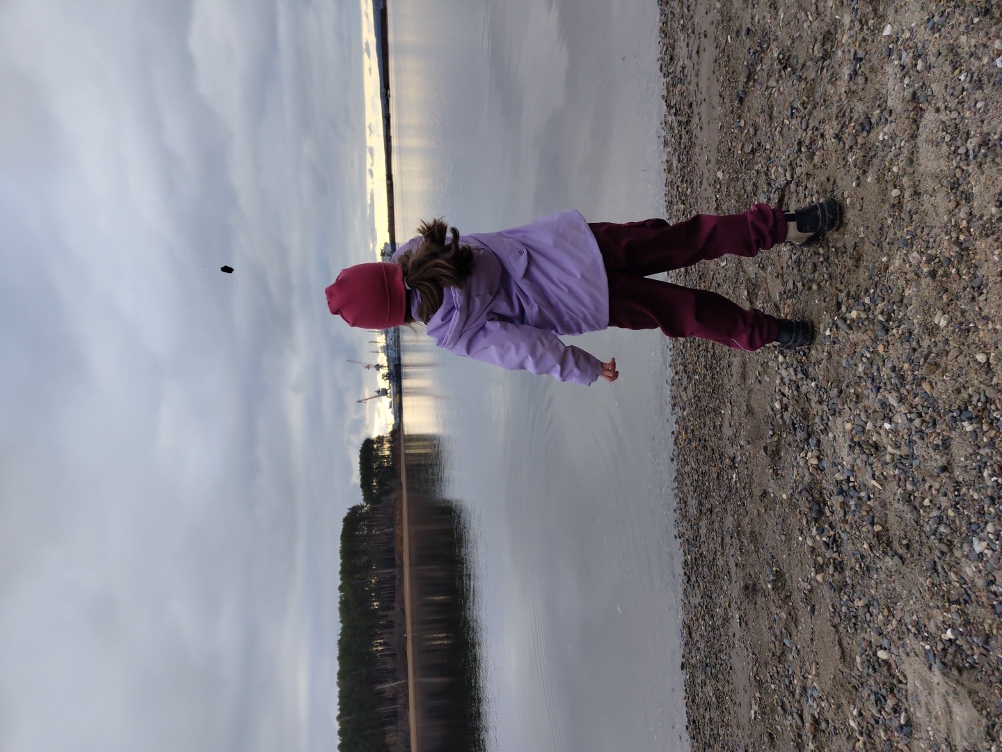 A child is captured from behind, standing on a pebbly and sandy shoreline next to a tranquil body of water under an overcast sky. The child, wearing a pink or reddish-purple beanie, a light purple jacket, and maroon pants, appears to have just thrown a small, dark pebble, which is visible in mid-air towards the center-left of the frame. Long hair in a ponytail is visible beneath the beanie.
The calm water, likely a large river or lake, perfectly reflects the sky and distant features. Across the water, a dark, dense treeline, possibly coniferous, stretches horizontally, its reflection clear on the water's surface. Further along the opposite shore, industrial structures, including cranes, can be seen, also mirrored in the still water. The sky is predominantly overcast with various shades of grey, though subtle patches of brighter, possibly golden light are reflected on the distant horizon, suggesting either an early morning or late afternoon light attempting to penetrate the clouds.
The immediate foreground shows the texture of the pebbly and sandy beach. No text is visible in the image. The scene conveys a quiet, perhaps contemplative, moment of play by the water, contrasting a natural shoreline with distant industrial elements.