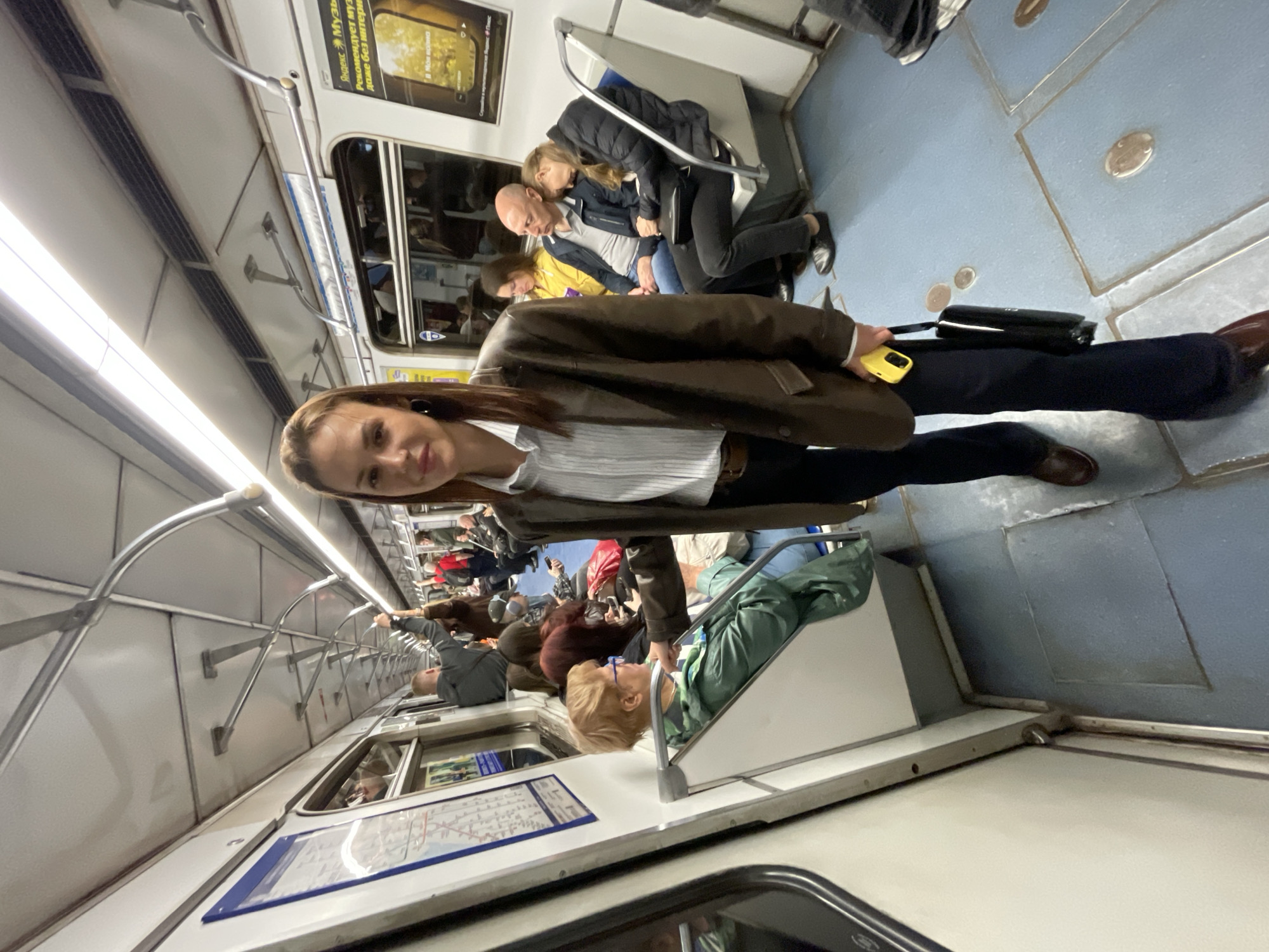The image shows the interior of a train car, likely a subway or metro, in an urban environment. The car is occupied by multiple people, most of whom are either seated or standing. The composition features a woman in the foreground wearing a brown jacket and holding a yellow mobile phone, with her back turned towards the camera while turning to the side. She also carries a black bag. Several other passengers are visible, including a couple seated with one leaning on the other, and an older woman seated with a green blanket. The car's interior is visible with advertising panels on the wall and a metal railing for passengers to hold onto. The lighting is bright. The overall scene suggests a daily commute in a busy city.