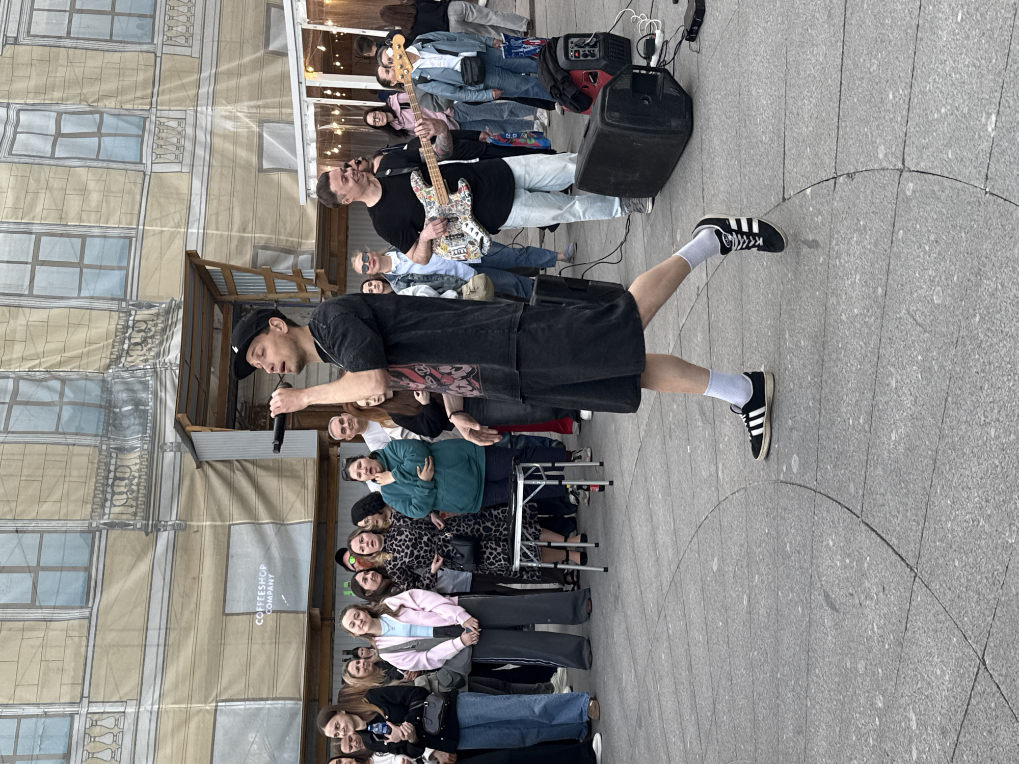 This is a medium shot of an outdoor street performance in Saint Petersburg, Russia. A male singer, wearing a black cap and shorts, is holding a microphone and singing to a crowd of onlookers. To his right, a male guitarist, wearing a black t-shirt and light-colored pants, is playing a white electric guitar covered in stickers.
In the background, a large mural depicts a classical building facade with windows and balconies. A temporary structure with a canopy is also visible, with string lights hanging from it. The crowd consists of men and women of various ages, some holding drinks and others watching the performance. The ground is paved with grey square tiles. Two large black speaker boxes and a portable power generator are placed on the ground to the right of the performers.