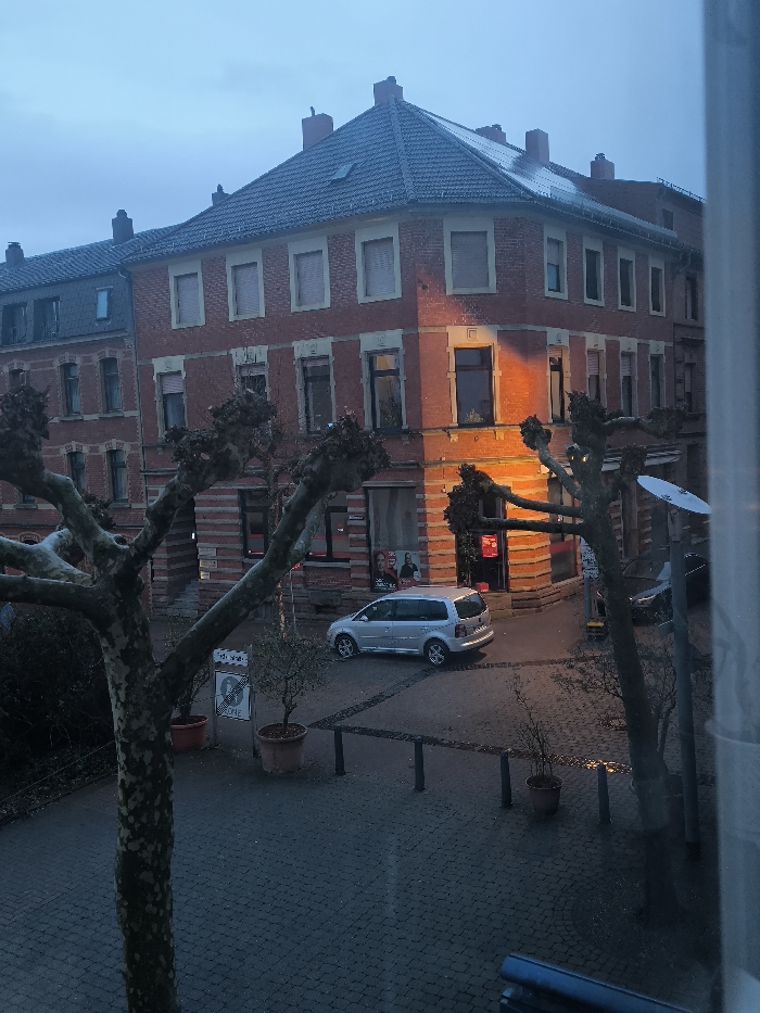 The image captures an elevated view of a street scene in Neunkirchen, Germany, under overcast conditions, likely at dusk or dawn. The central focus is a three-story red brick corner building with a dark tiled roof and multiple windows, some fitted with light-colored external blinds. A prominent orange light source illuminates a corner window and adjacent wall of this building. The ground floor of the corner building features commercial spaces with large display windows, one containing a poster depicting two individuals, and another faintly revealing a person inside behind a parked vehicle.
To the left, an adjacent multi-story red brick building with a dark roof continues the urban architectural style. The street below is paved with dark cobblestones, appearing damp or wet, reflecting ambient light. A silver Volkswagen minivan, likely a Touran, is parked on the cobblestone street near the corner building. The front section of a dark vehicle is partially visible on the far right.
Several large, heavily pruned deciduous trees with bare, gnarled branches dominate the foreground and frame the view. Potted plants are positioned on the cobblestone sidewalk. Black bollards define the edge of the pedestrian area, and a white traffic sign indicating a "ZONE" is visible. A white satellite dish is mounted on a pole or tree structure on the right side of the image. The sky is a uniform, light blue-grey. A window frame is partially visible on the extreme right, indicating the elevated vantage point.