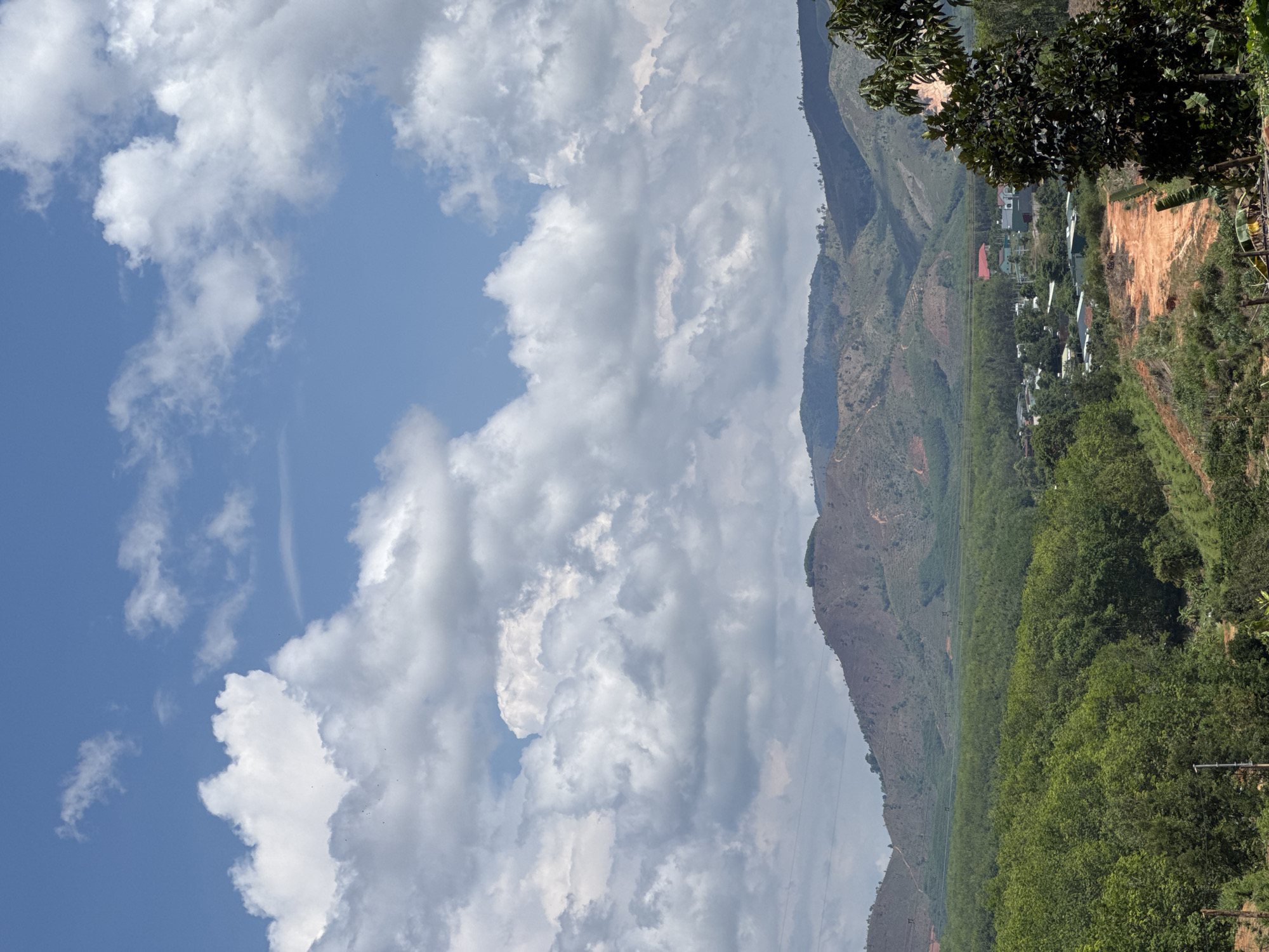 A landscape photograph captures a view in Bờ Y, Vietnam. The foreground features lush green foliage, transitioning to a hillside with sparser vegetation. A power line is visible. The background is dominated by a large expanse of sky filled with fluffy white clouds, with patches of blue sky breaking through. A few structures are present among the vegetation.