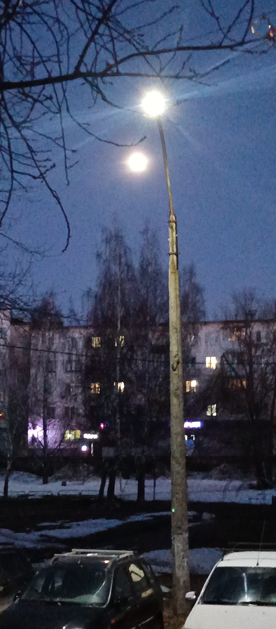 The image depicts an urban scene at dusk or night. A tall, weathered concrete lamppost stands prominently in the foreground, topped with a bright light fixture. A second, dimmer light source appears higher in the sky, potentially the moon. Leafless tree branches frame the top left corner of the image.
Behind the lamppost, a multi-story residential building is visible, with several illuminated windows suggesting occupancy. The building is partially obscured by bare trees. A layer of snow covers the ground in the foreground, and parked cars are visible along the bottom edge of the frame. The sky is a dark, muted blue.